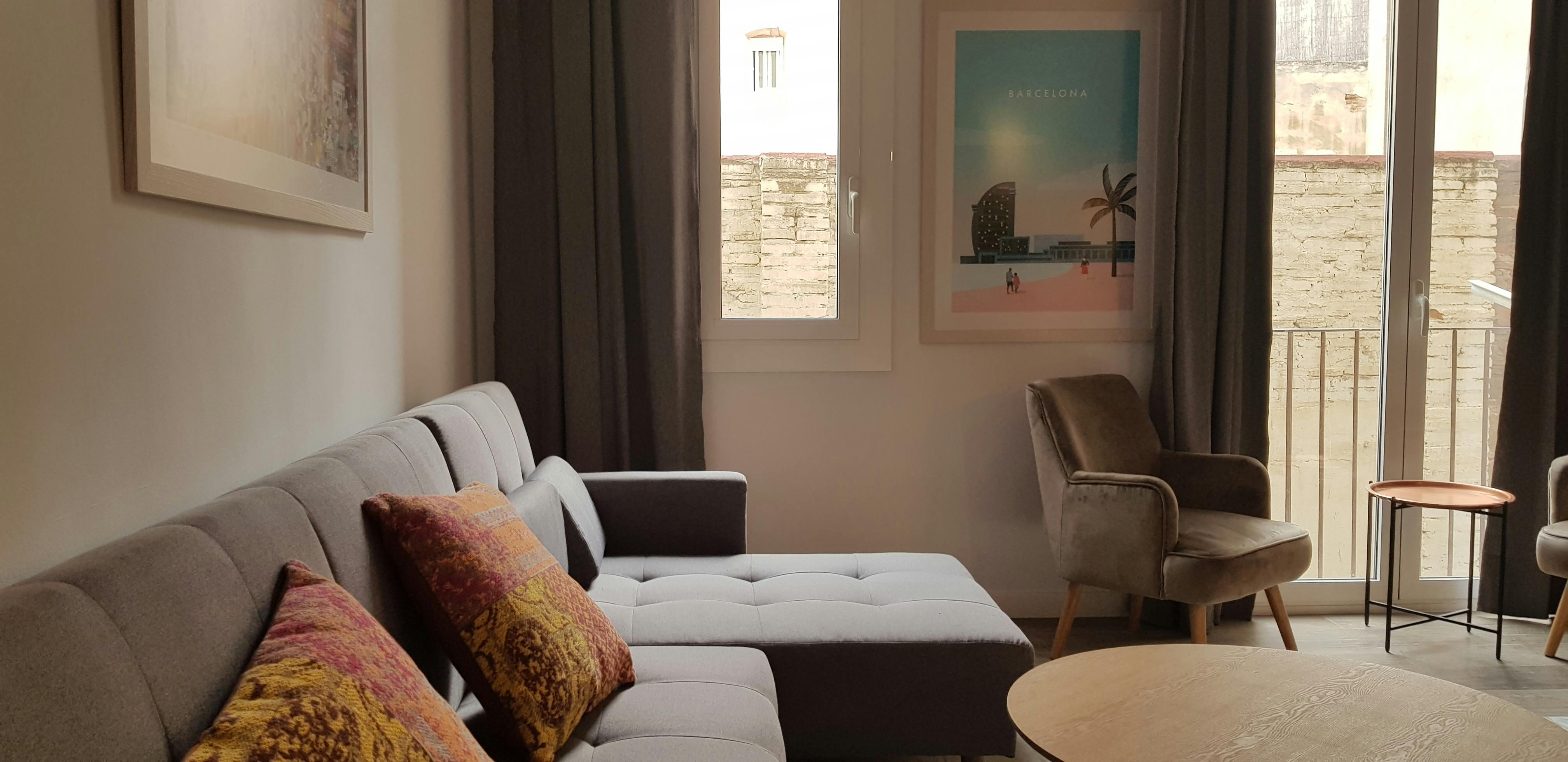 The living room features a comfortable-looking grey sofa with pillows, a large window with curtains. Natural light floods the space, illuminating the wooden coffee table and a nearby armchair.