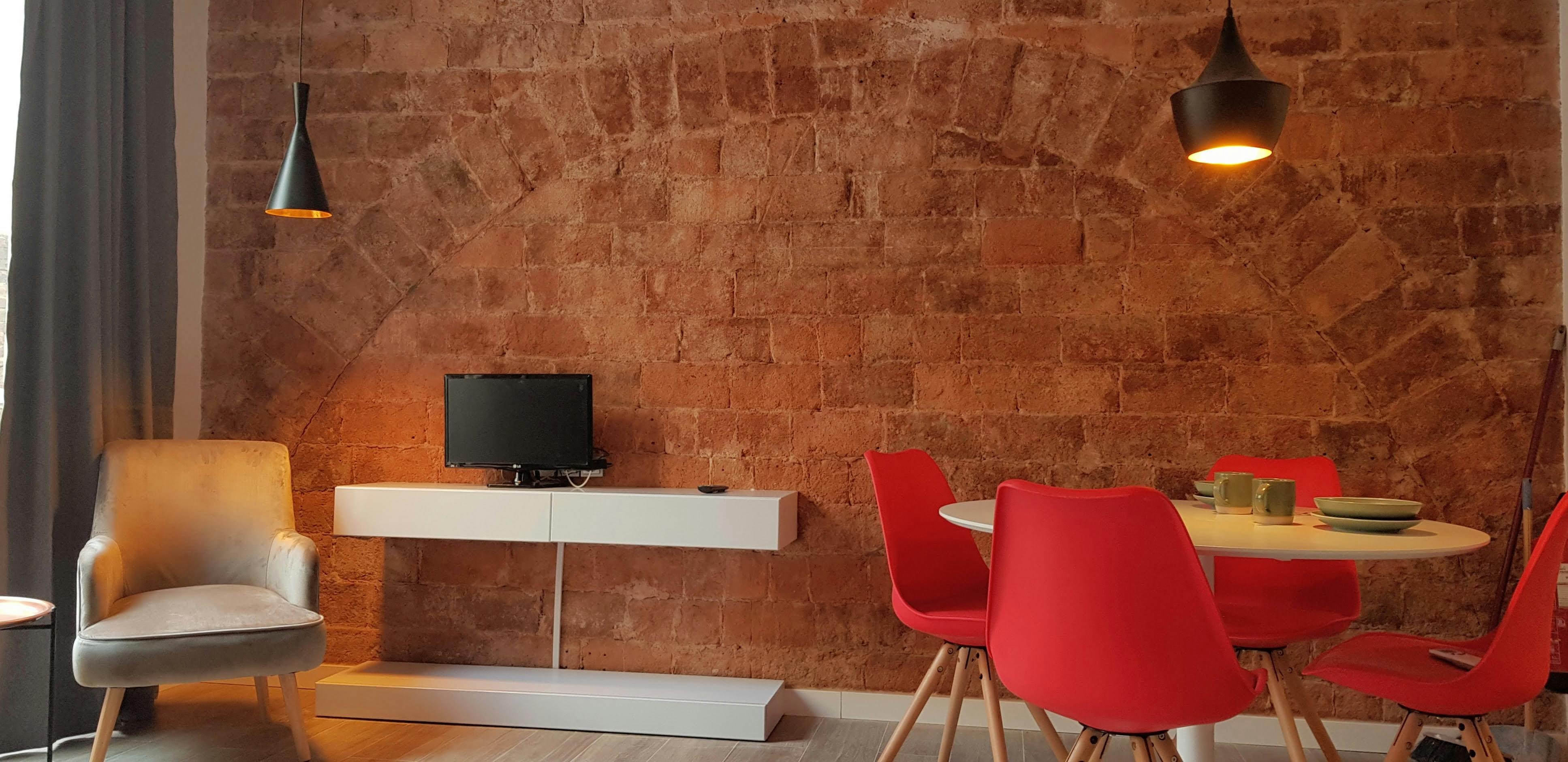 The living room features a modern design with exposed brick walls, a flat-screen television, and a dining table with four bright red chairs. Natural light floods the space, highlighting the exposed brickwork and creating a warm and inviting ambiance.
