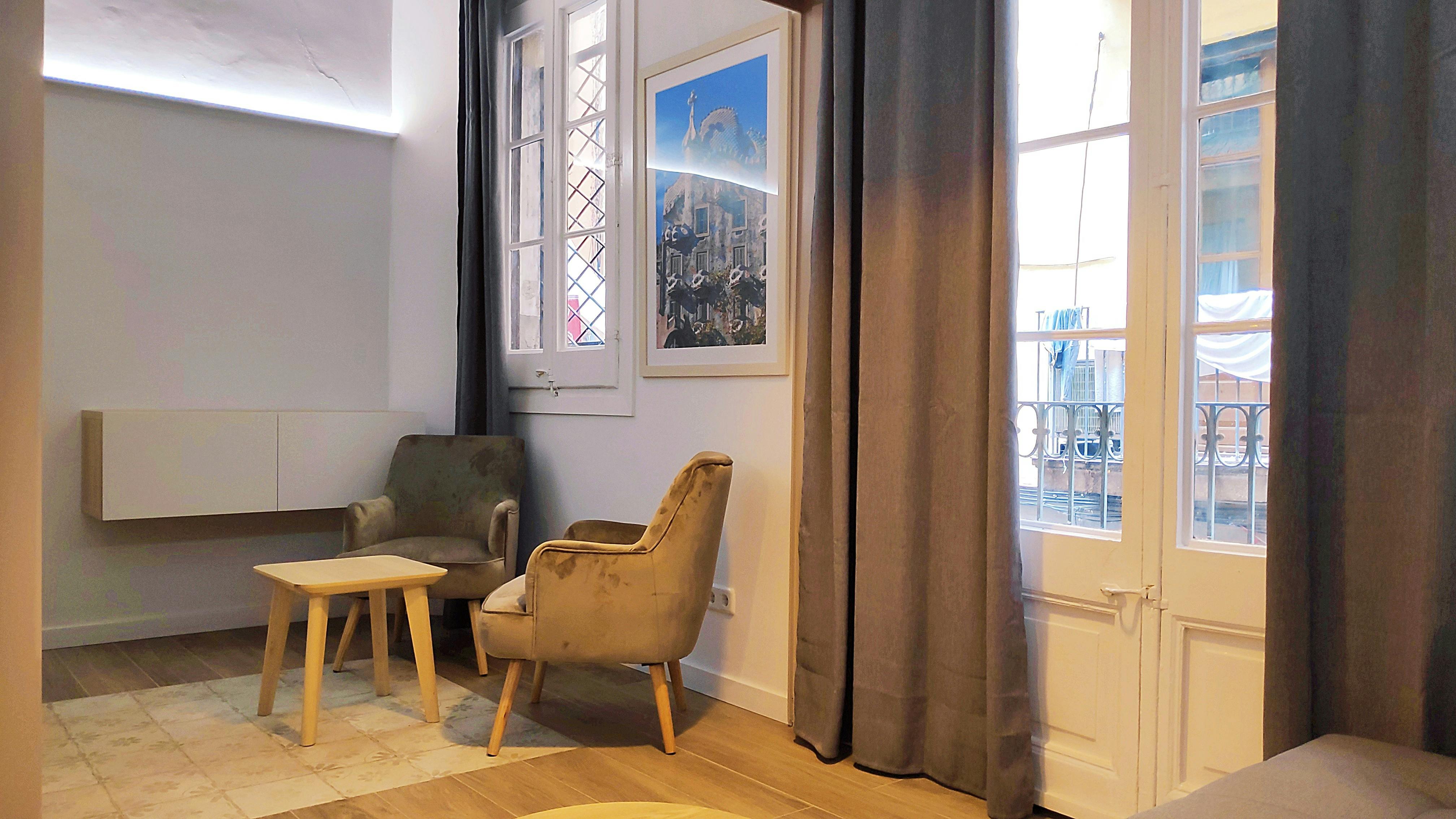 The living room features two plush armchairs, perfect for relaxing after a day exploring the city. A small, wooden table sits between the chairs. The room is bathed in natural light from the large window with a small balcony.