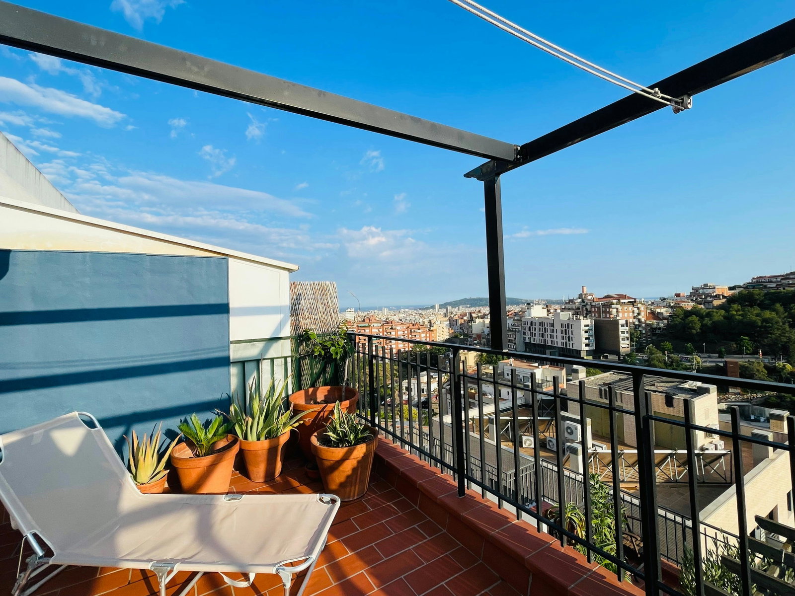 Enjoy breathtaking views of the Barcelona skyline from the comfort of this private terrace, complete with a lounge chair and potted plants. The terrace features a pergola for shade and a black metal railing.