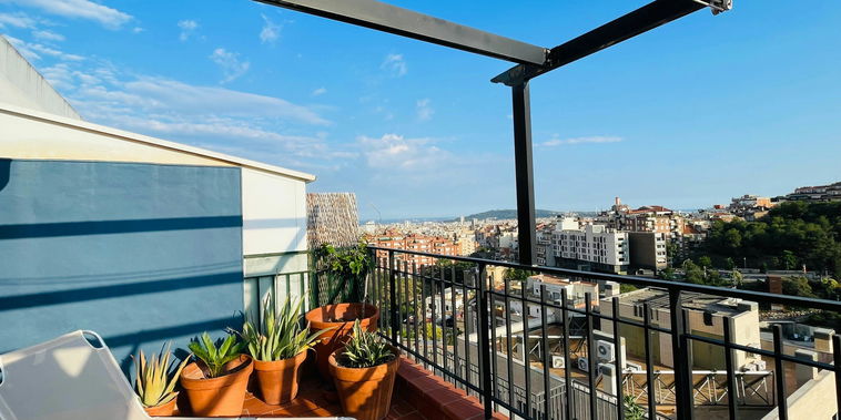 Enjoy breathtaking views of the Barcelona skyline from the comfort of this private terrace, complete with a lounge chair and potted plants. The terrace features a pergola for shade and a black metal railing.