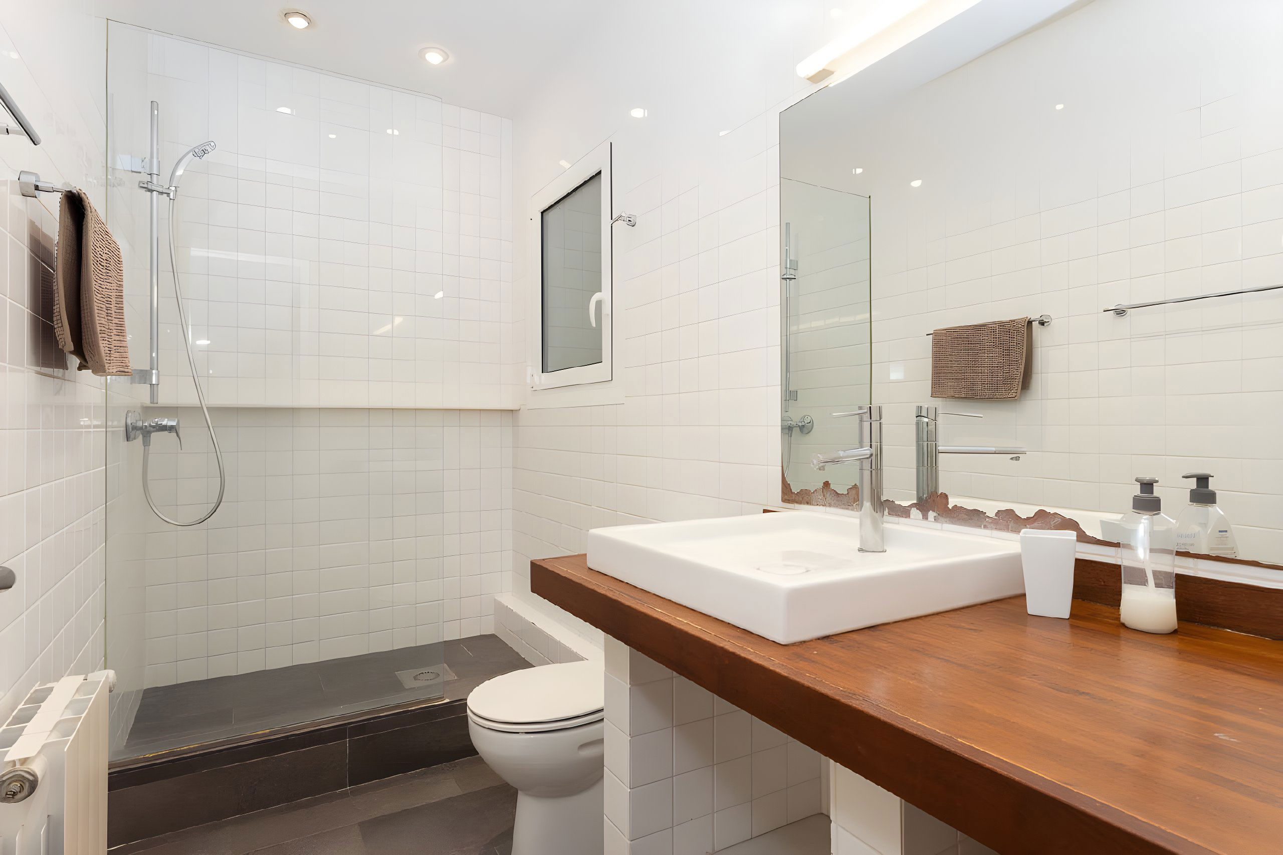 The bathroom features a shower stall with a glass door, a white porcelain toilet, and a modern sink with a sleek chrome faucet. The countertop is made of rich, dark wood, providing a warm contrast to the white tiled walls. There is a mirror that extends the length of the counter, reflecting the clean lines of the room. Small window.