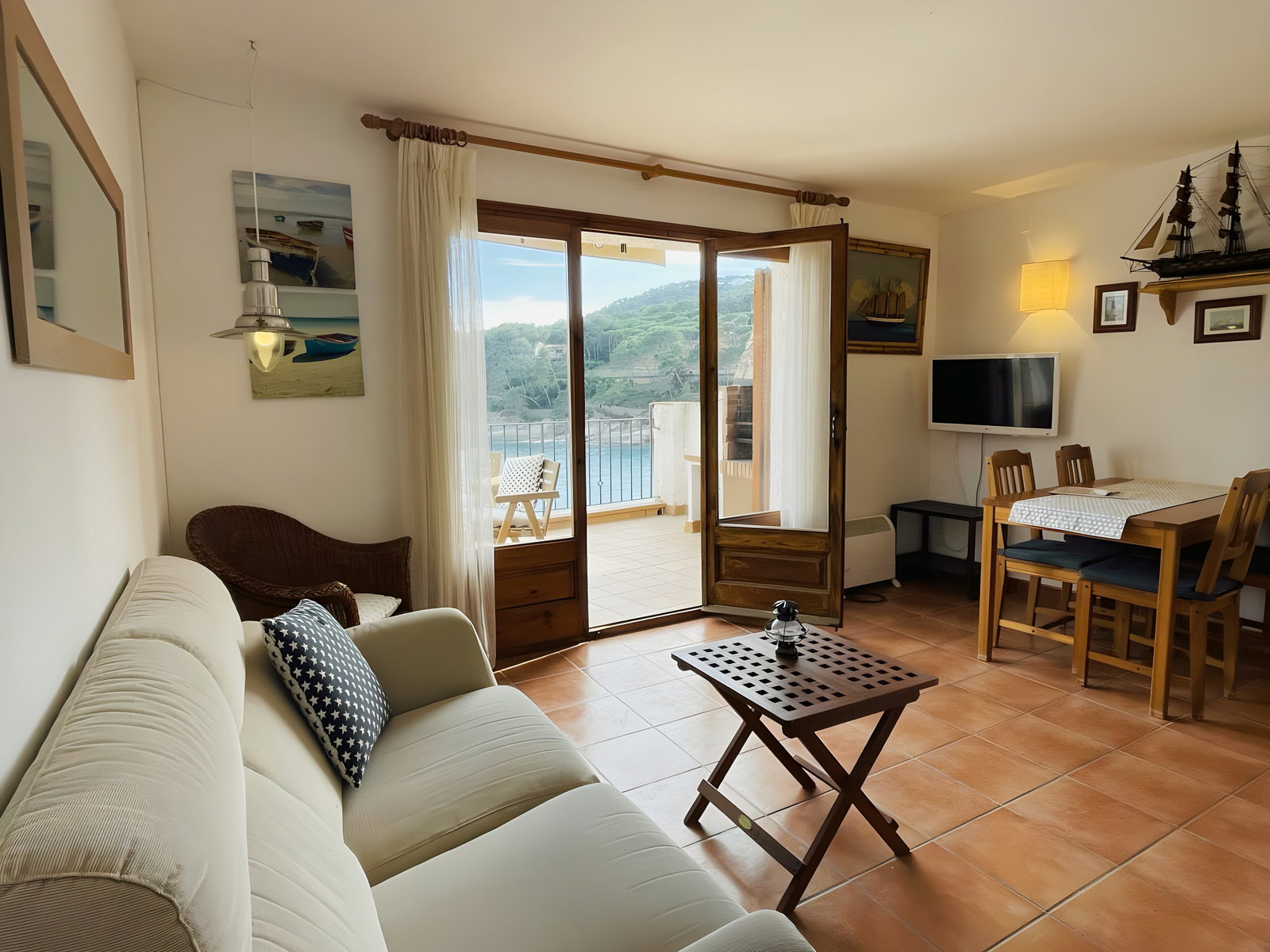 This is a view of the spacious living room, featuring a comfortable white sofa, a wooden table with four chairs, and a flat-screen TV. The large patio doors open to a private terrace overlooking the sea, providing stunning panoramic views of the coastline. The tiled floor adds a warm and inviting touch to the overall design, creating a relaxed and airy atmosphere.