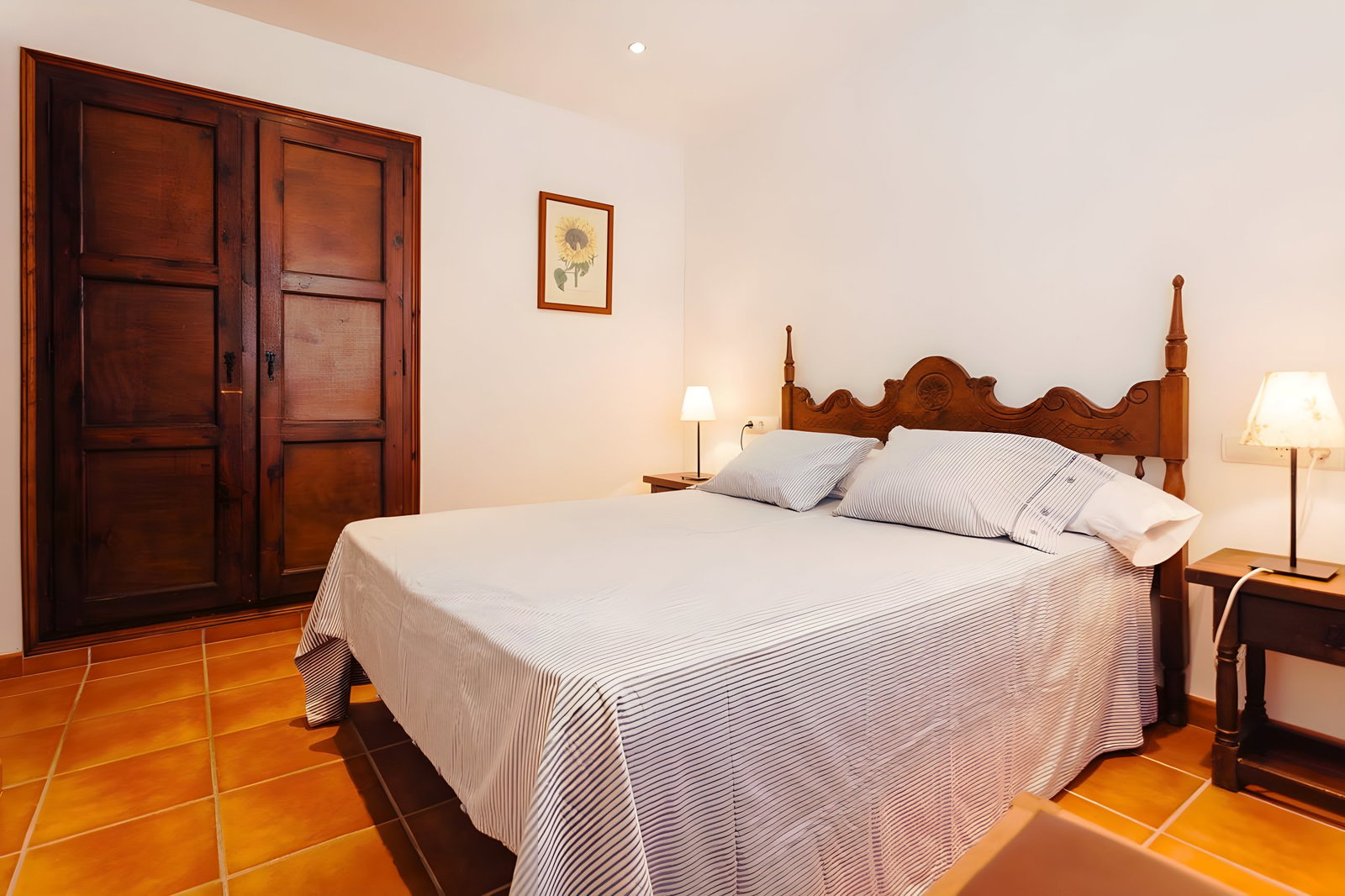This bedroom features a queen-size bed with a striped duvet and two matching pillows. The headboard is a beautifully carved piece of furniture that adds a touch of elegance to the room. The tiled floors and wooden closet doors create a warm and inviting atmosphere, while the picture of a sunflower above the closet adds a cheerful touch. This room is sure to provide a restful and comfortable stay for guests.