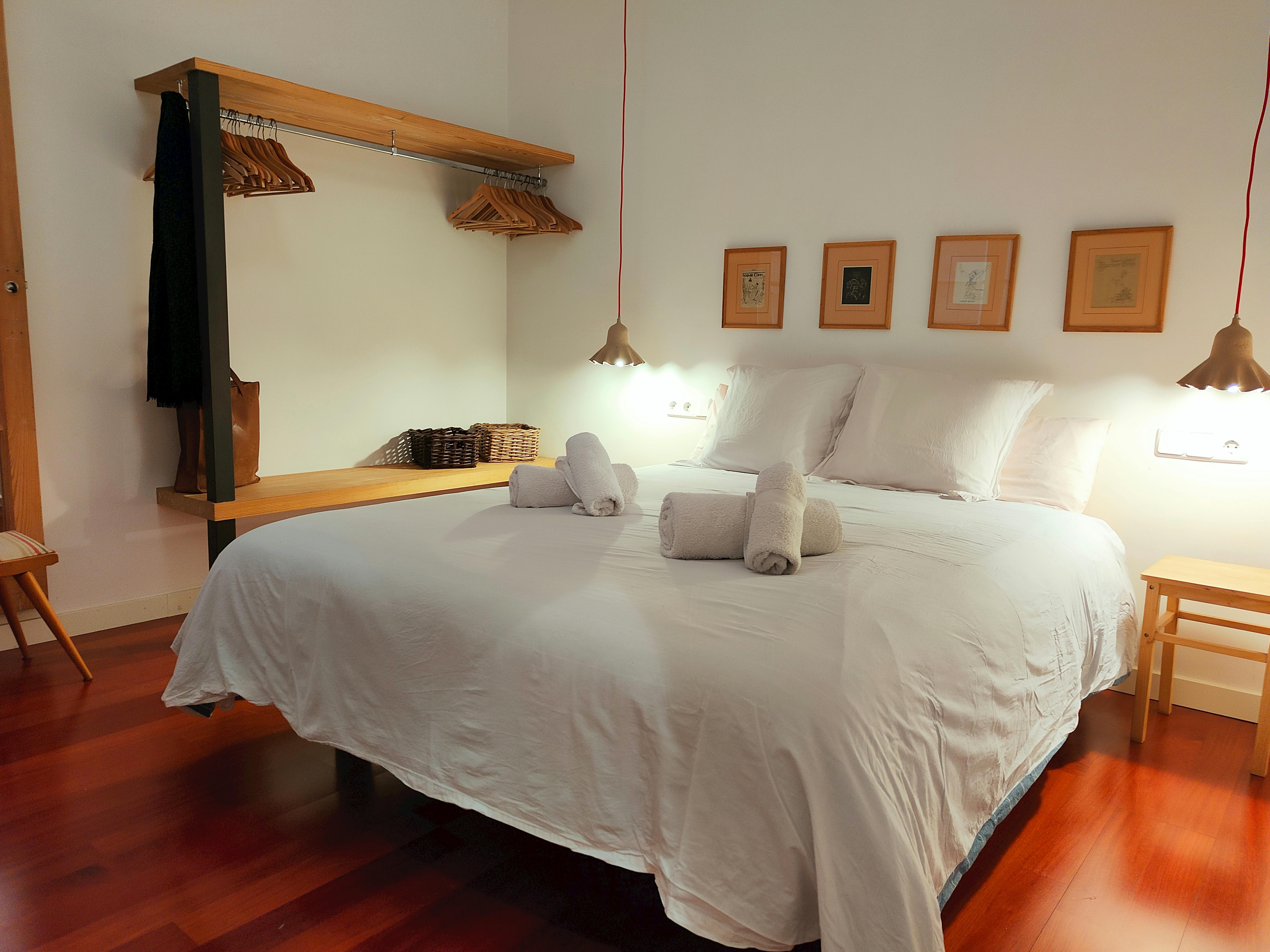 This bright bedroom features a double bed, a wooden floor, and an open closet. The room is decorated with framed artwork, a small bedside table, and a bedside lamp. The room is spacious offering a comfortable and inviting space for guests.