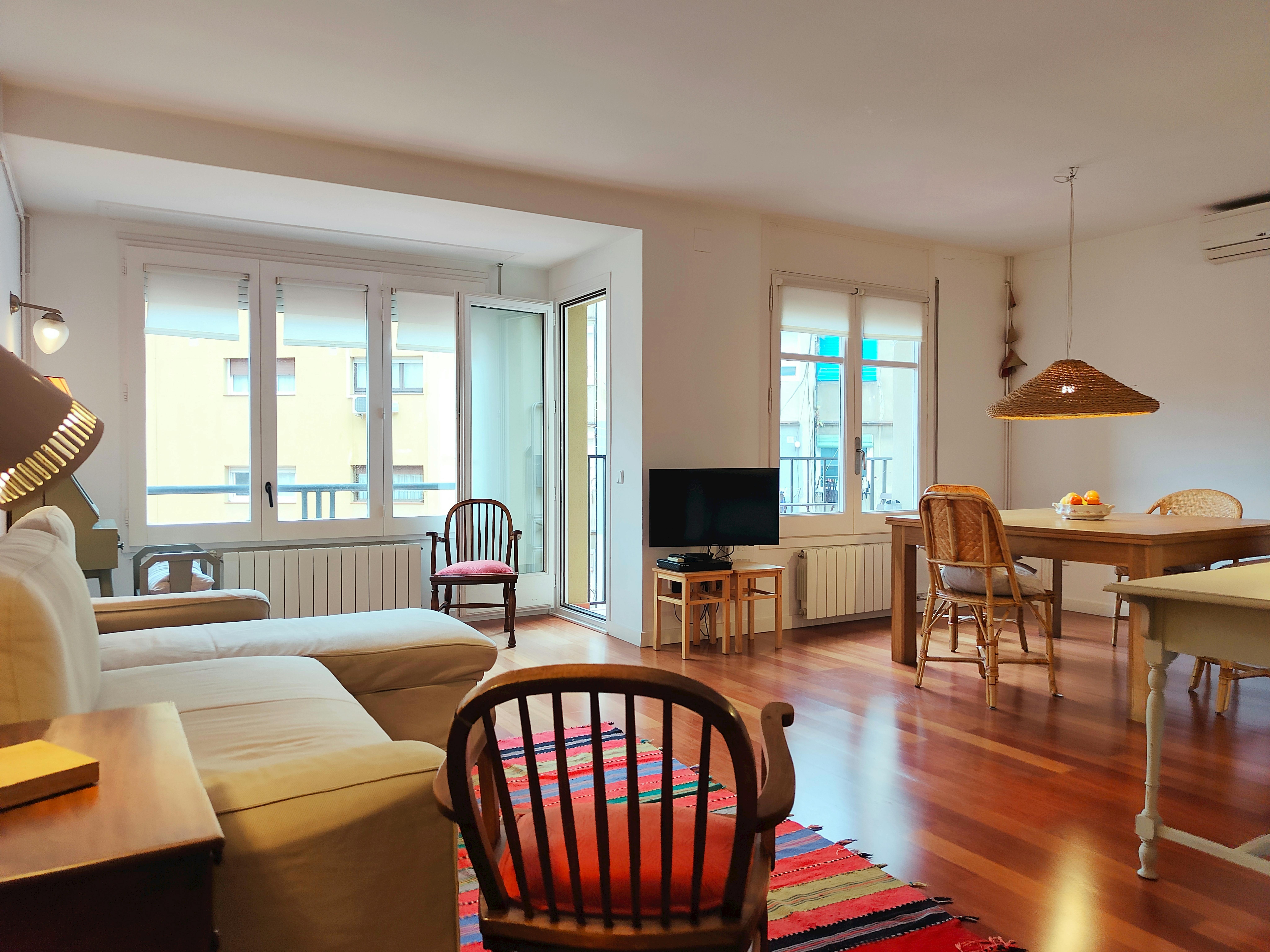 The spacious living room of the apartment is bathed in natural light streaming through large windows, showcasing the warm hardwood floors and a comfortable white couch with pillows. The living room features a large flat-screen TV and a dining table in the background.