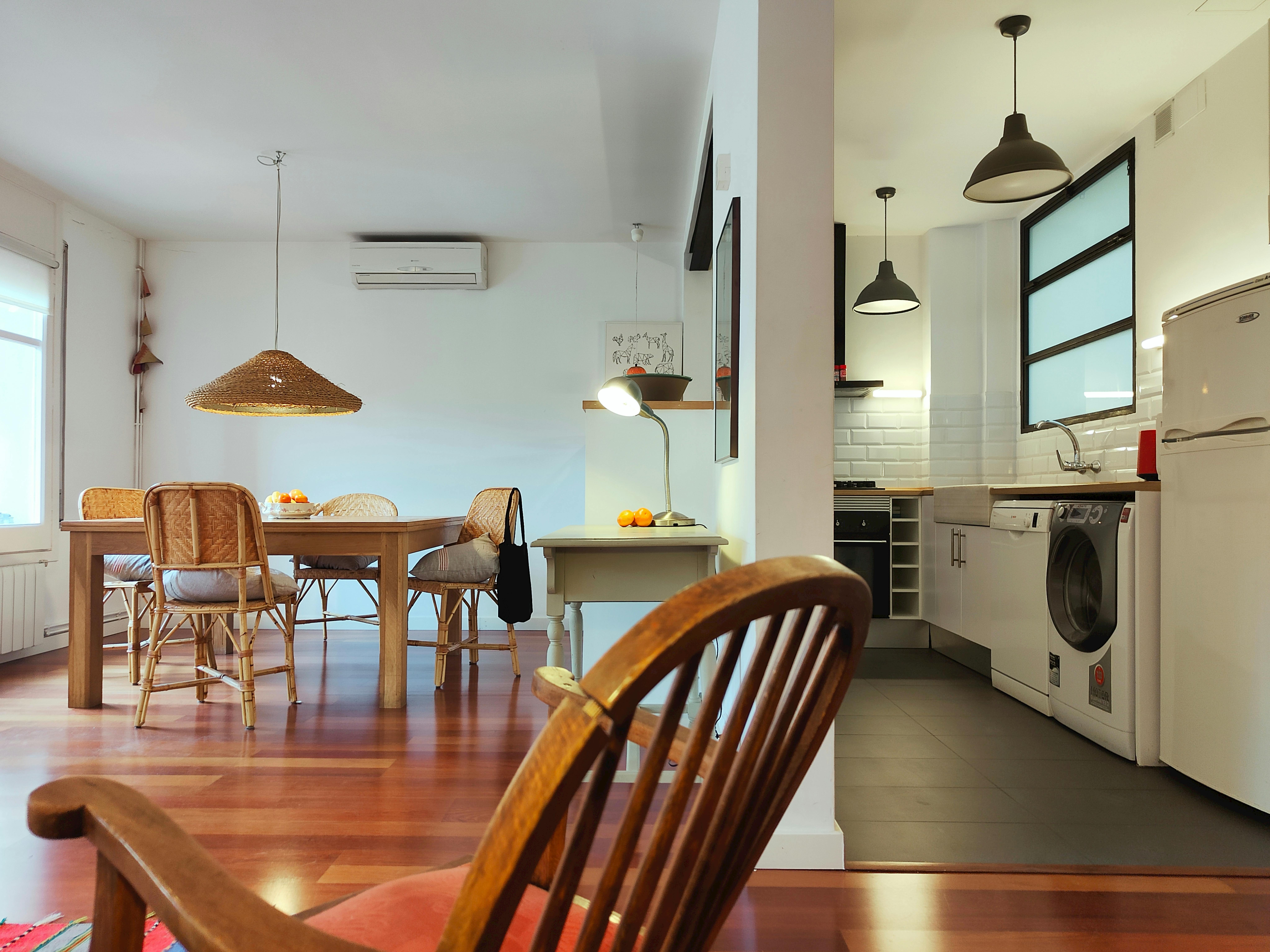 The spacious living area features a large wooden dining table with wicker chairs, a unique woven pendant light, and a view into the open kitchen. The kitchen is equipped with modern appliances, including a dishwasher, washing machine, and a sleek stovetop. The apartment boasts warm hardwood floors throughout and is tastefully decorated with a mix of modern and traditional elements.