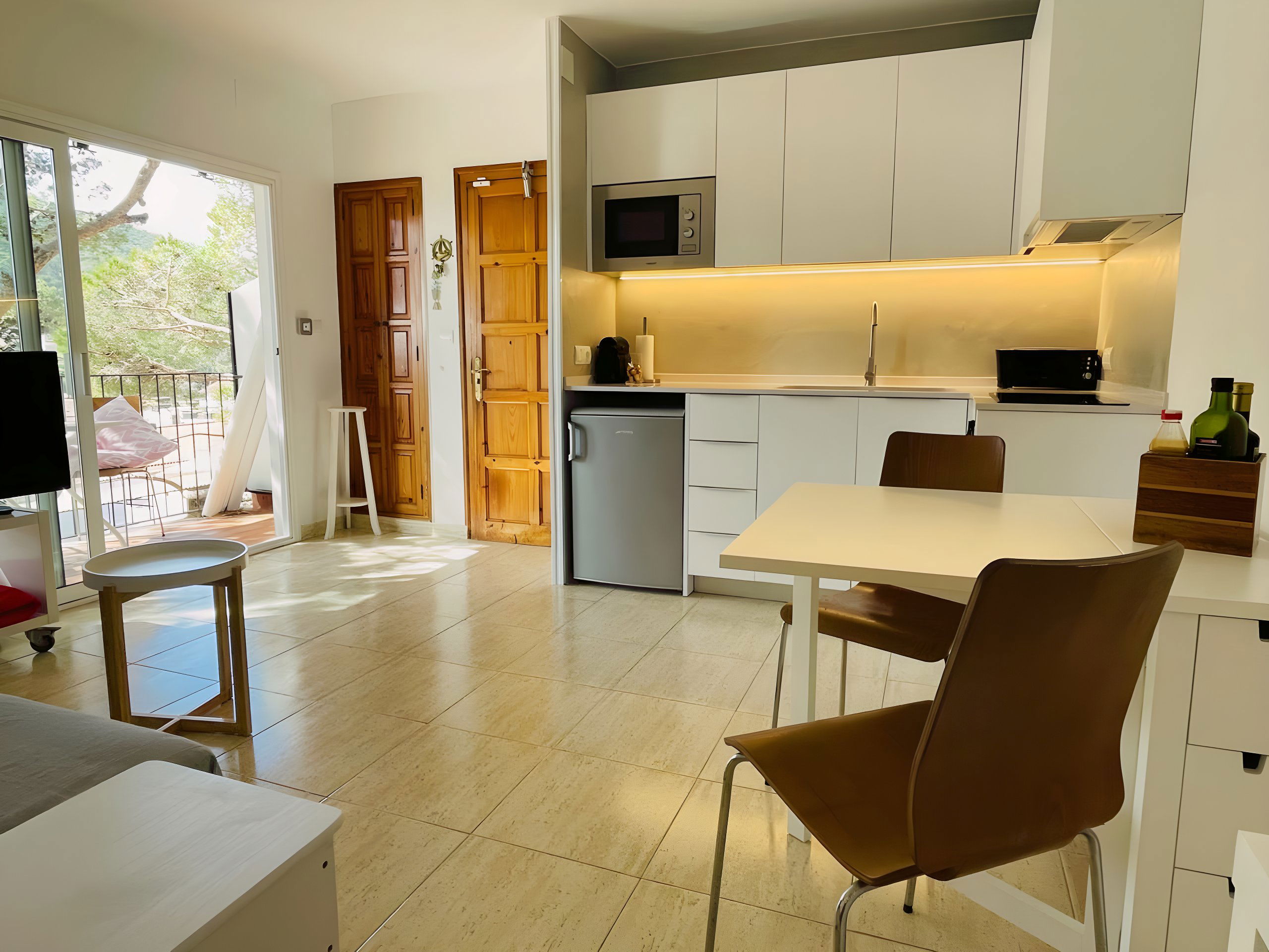 The bright and airy living space features a modern, fully equipped kitchen with white cabinets and a sleek countertop, perfect for preparing delicious meals during your stay. The tiled floors add a touch of elegance, while the sliding glass doors offer breathtaking views of the lush greenery outside, leading to a private balcony.