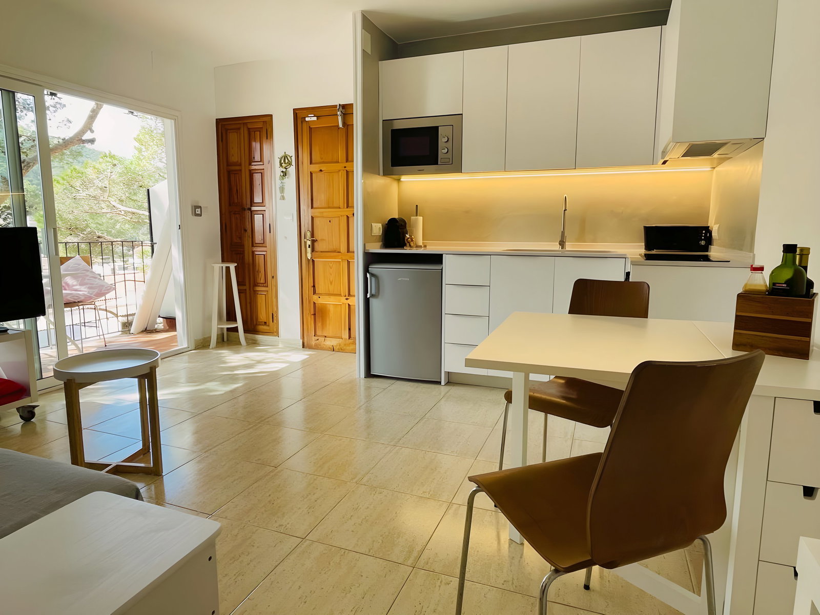 The bright and airy living space features a modern, fully equipped kitchen with white cabinets and a sleek countertop, perfect for preparing delicious meals during your stay. The tiled floors add a touch of elegance, while the sliding glass doors offer breathtaking views of the lush greenery outside, leading to a private balcony.