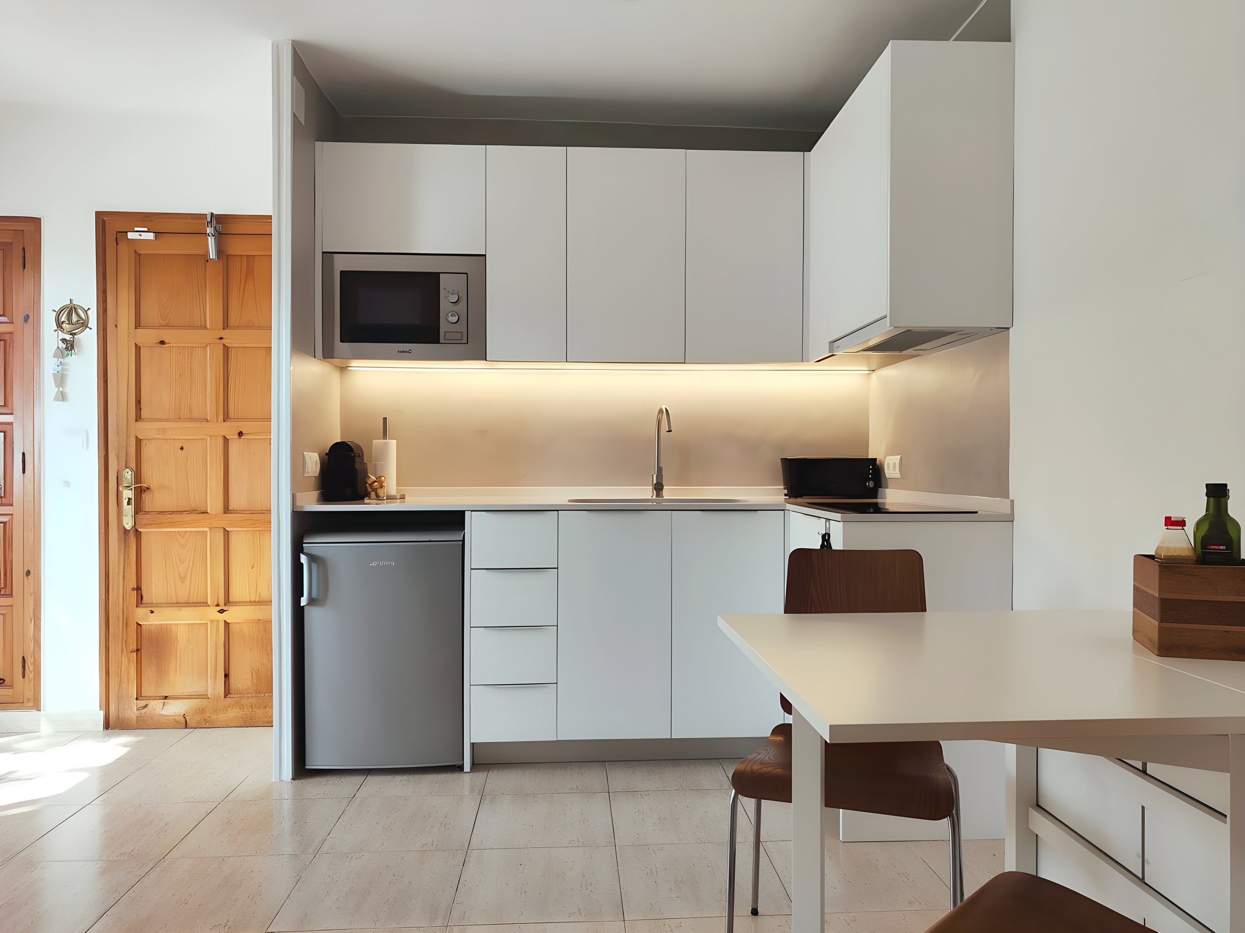 The bright and modern kitchen boasts sleek white cabinets and a compact layout. A small refrigerator, microwave, and coffee machine offer convenience for preparing meals, while a dining table and chairs provide a comfortable space for enjoying them. Natural light pours in through a nearby door, illuminating the tiled floor. This is a  functional and stylish kitchen.