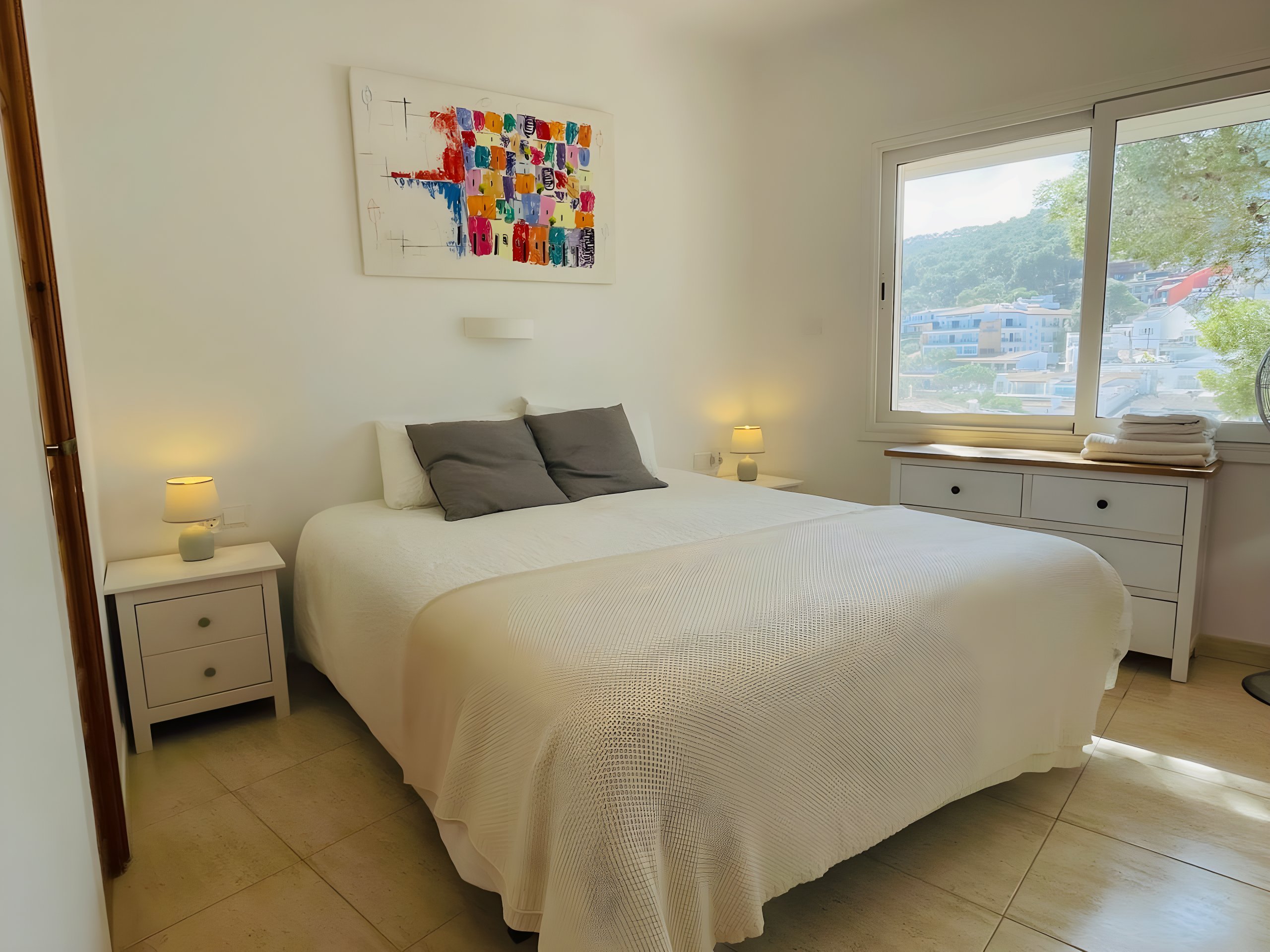 The bedroom features a queen-sized bed with crisp white linens and two plush gray throw pillows.  A large window offers stunning views of the surrounding hills, while a colorful painting adds a touch of vibrancy to the space. The room is bathed in natural light, making it a peaceful and inviting retreat.