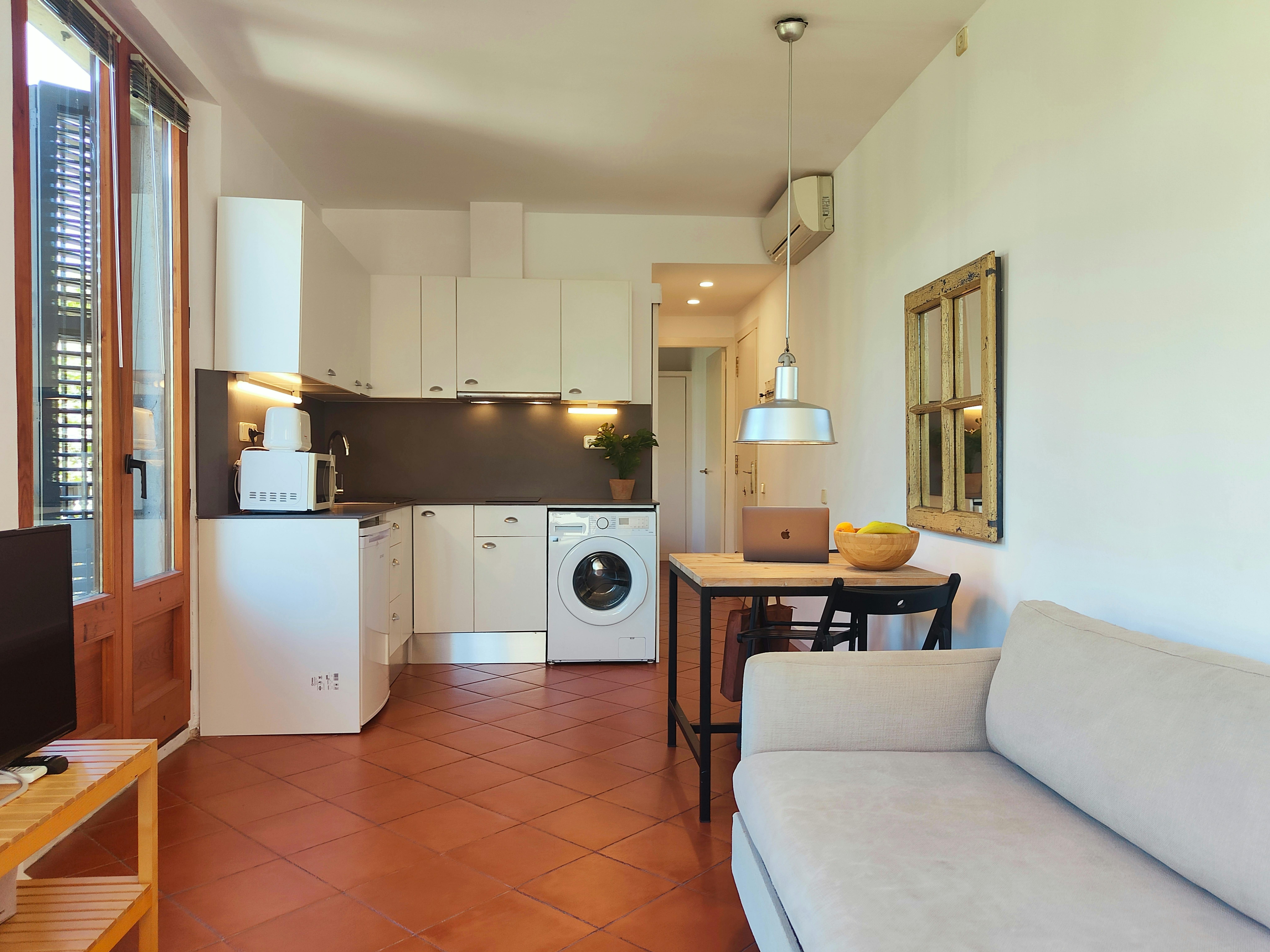 The living room features a comfortable sofa positioned in front of a wooden table. The apartment has terracotta tile floors throughout.