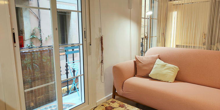 Natural light streams into this trendy Barcelona loft, illuminating a stylish living space designed for relaxation and urban exploration. The blend of modern furnishings and glimpses of traditional Spanish architecture through the balcony door creates a unique and inviting ambiance.
