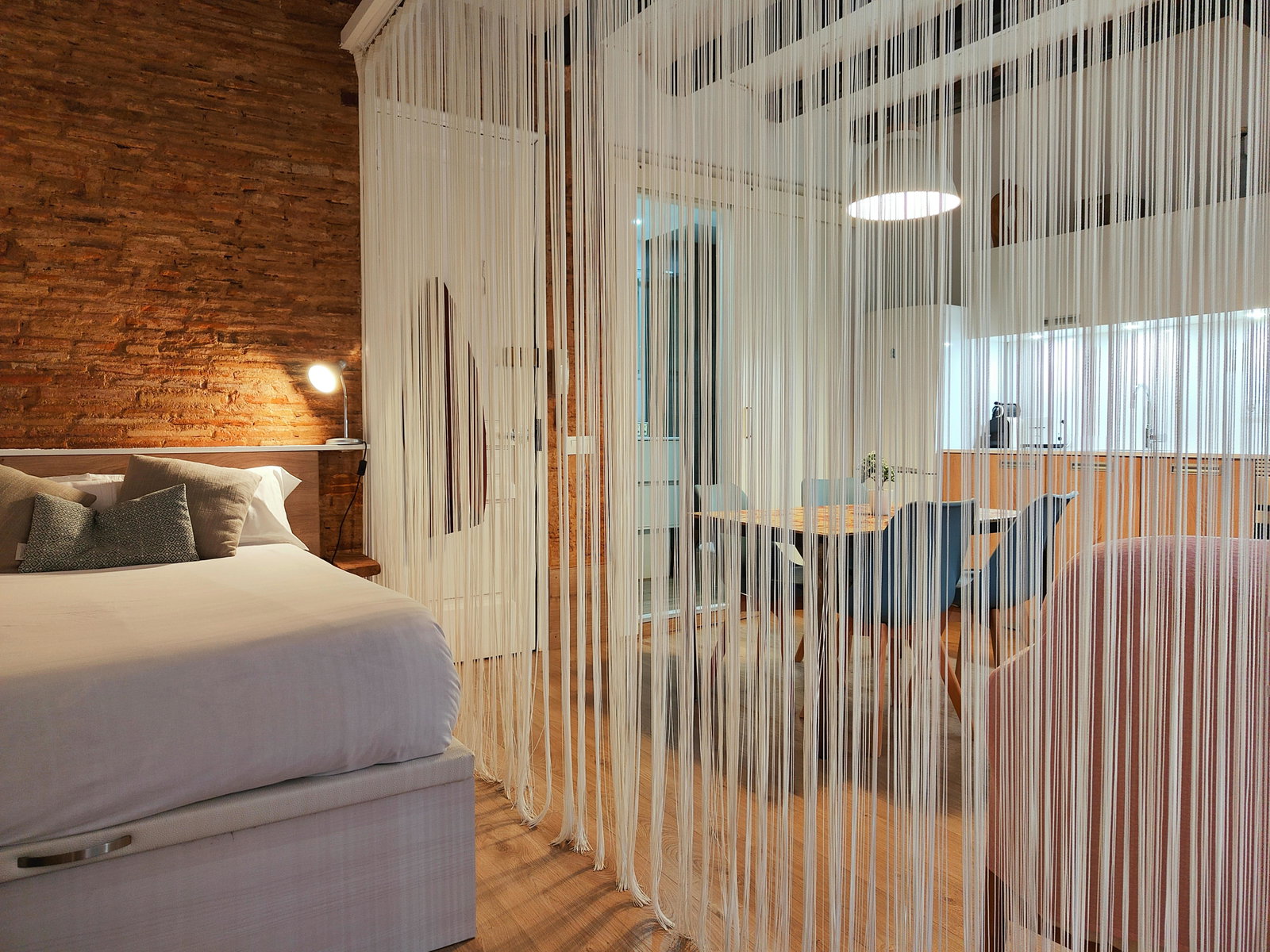The inviting bedroom features a comfortable queen-size bed with storage space underneath, perfectly positioned against a rustic brick wall. A unique string curtain separates the bedroom from the open kitchen and dining area, adding a touch of modern elegance to the loft. The kitchen is fully equipped with modern appliances, ensuring a comfortable and convenient stay for guests.  The wooden floor throughout the loft creates a warm and inviting ambiance, making it an ideal space for relaxing after a day exploring Barcelona.