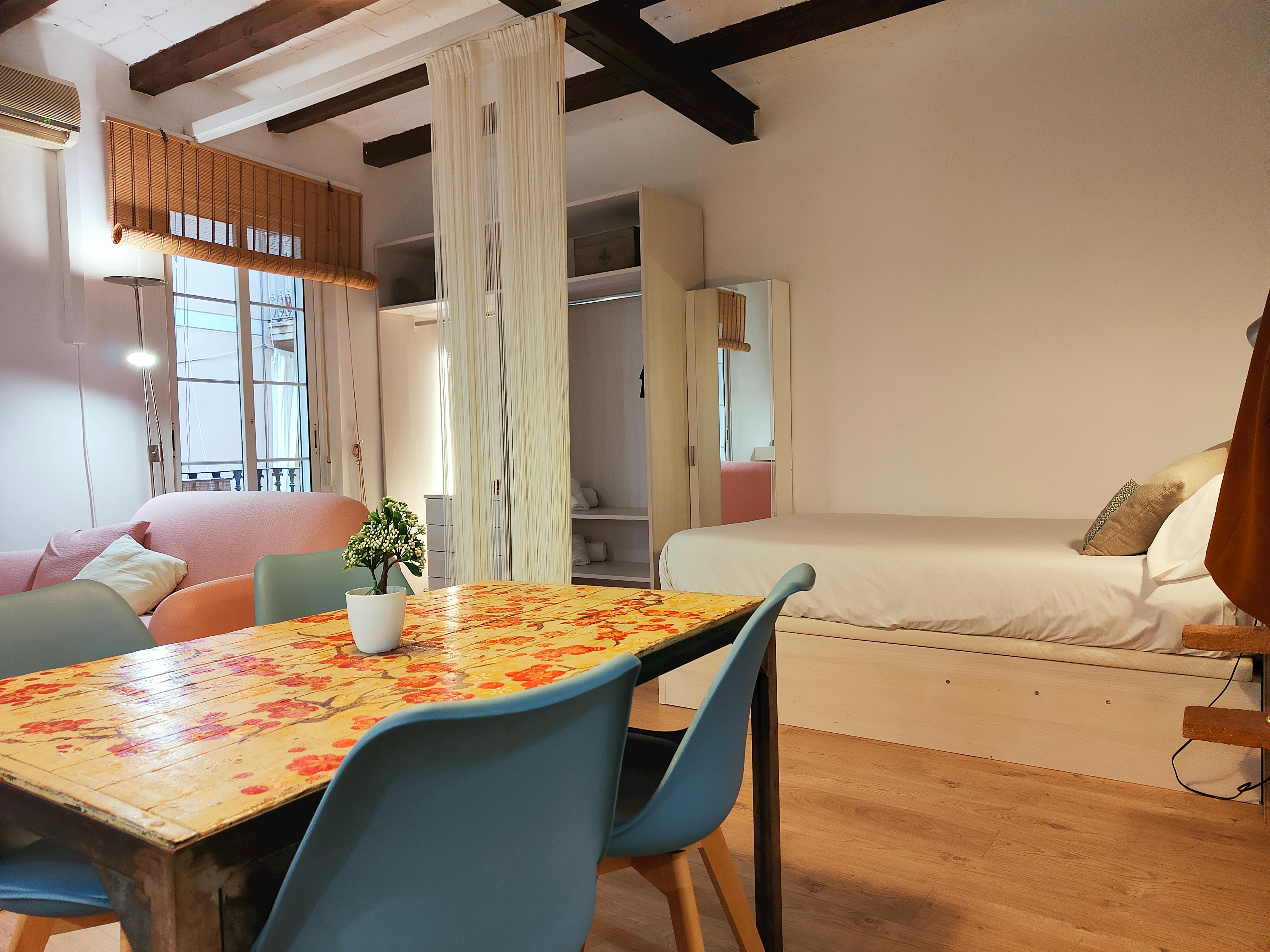 The spacious and well-lit living area features a dining table with four teal chairs, perfect for enjoying meals or working remotely. Open living & sleeping area with a queen size bed (with a storage place underneath it), a sofa, tv.