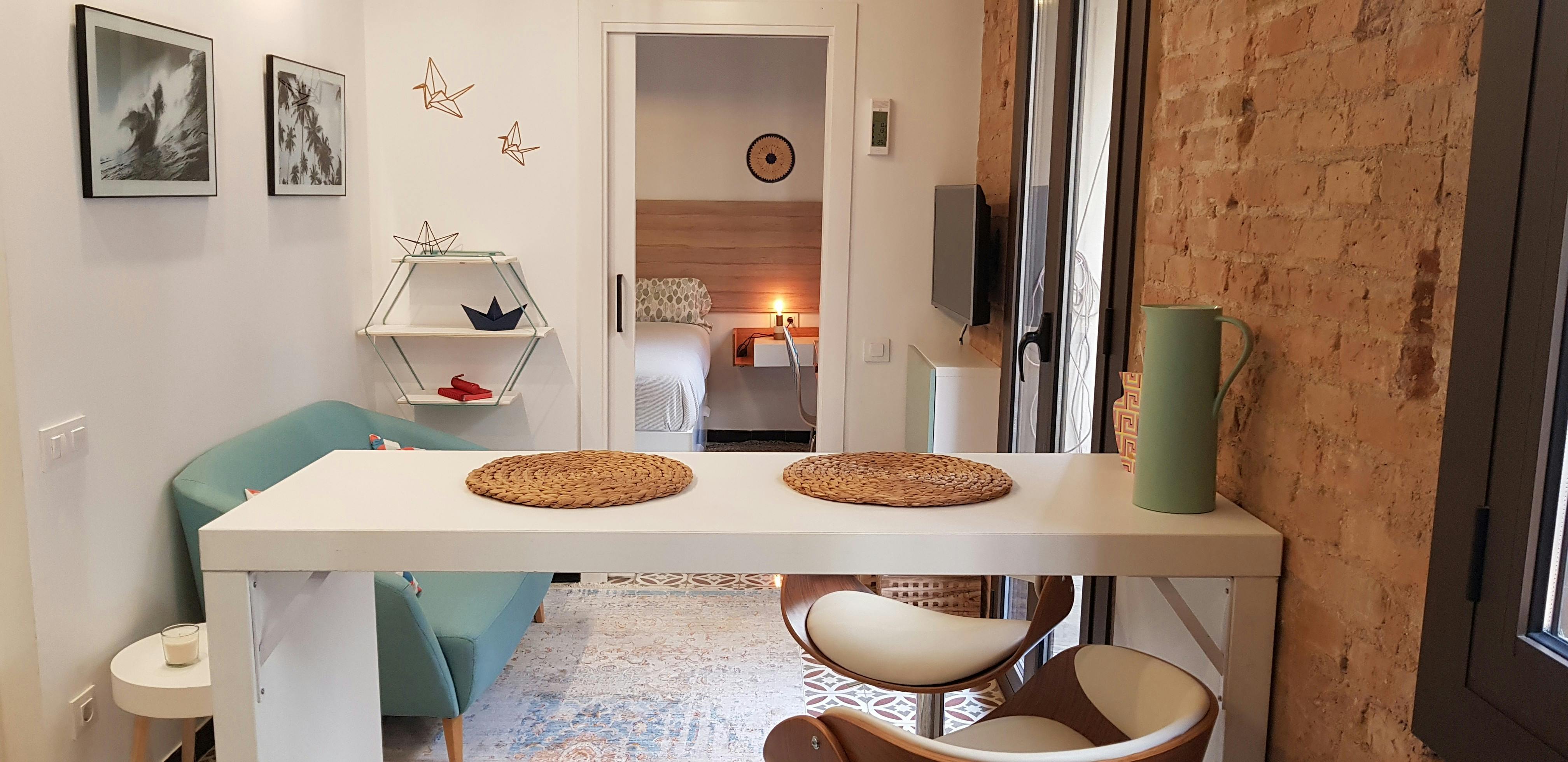 The open-plan living space features a white breakfast bar with two woven placemats and two white bar stools with wooden seats. Through the doorway, you can see a glimpse of the bedroom with a white bed and a wooden nightstand.  An exposed brick wall adds a touch of rustic charm to the space.  The space is well-lit by natural light streaming through the large window.