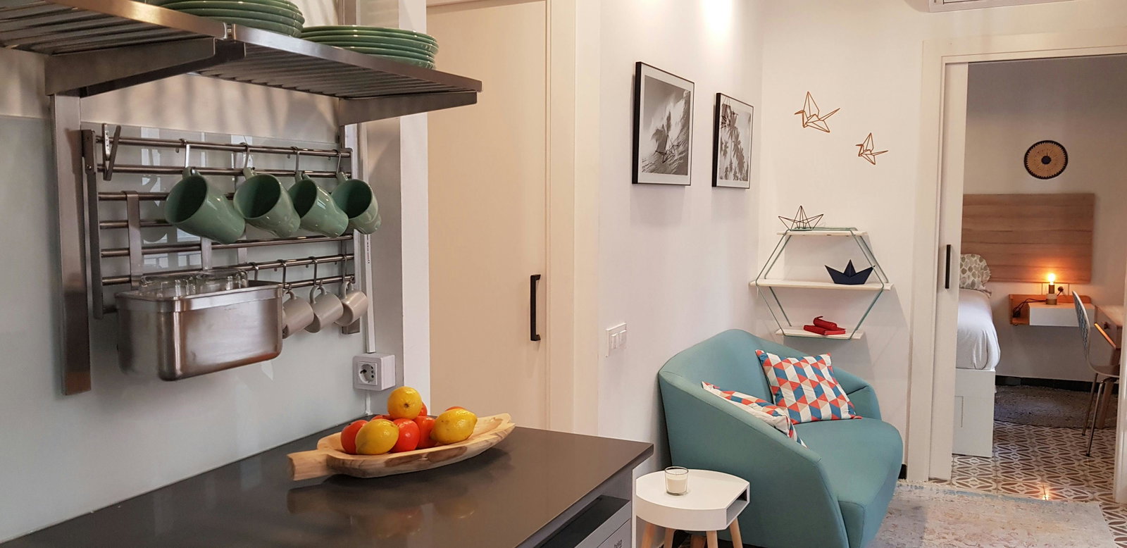 This open-plan living area features a stylish, teal sofa with geometric patterned cushions. The adjacent bedroom is visible through an open doorway, with a simple yet elegant wooden headboard and bedside lamp. This apartment is designed for those who value both style and functionality.