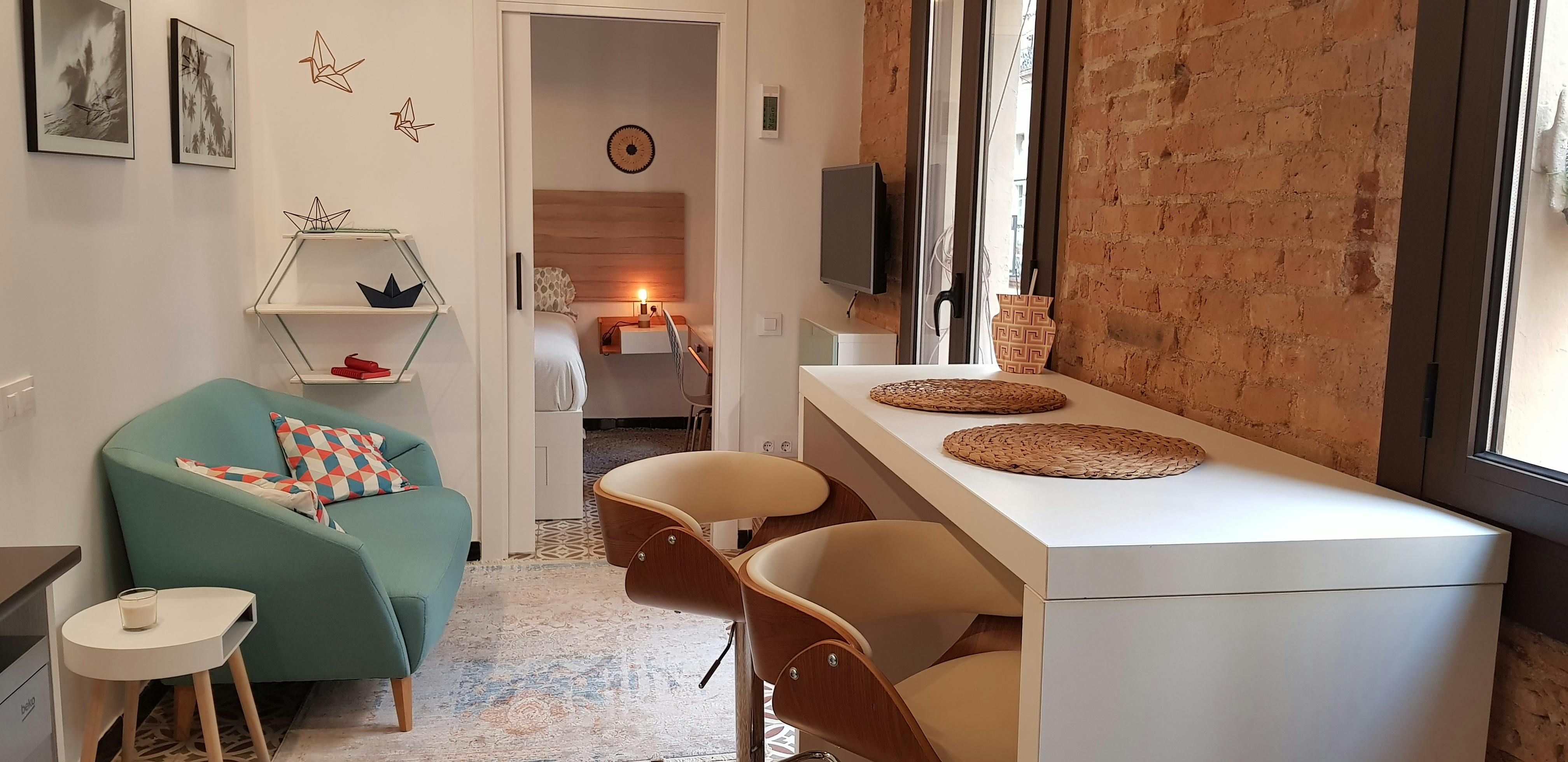 This is a view of the living room, featuring a light blue sofa with patterned cushions and a white side table with a candle. The room is tastefully decorated with minimalist furniture and an exposed brick wall.  A doorway leads into the bedroom, which features a queen-sized bed and a small desk.  The living room also includes a dining table with two chairs. There's also a window with a view of the city.  The overall ambiance of the room is bright and modern, with a touch of warmth.