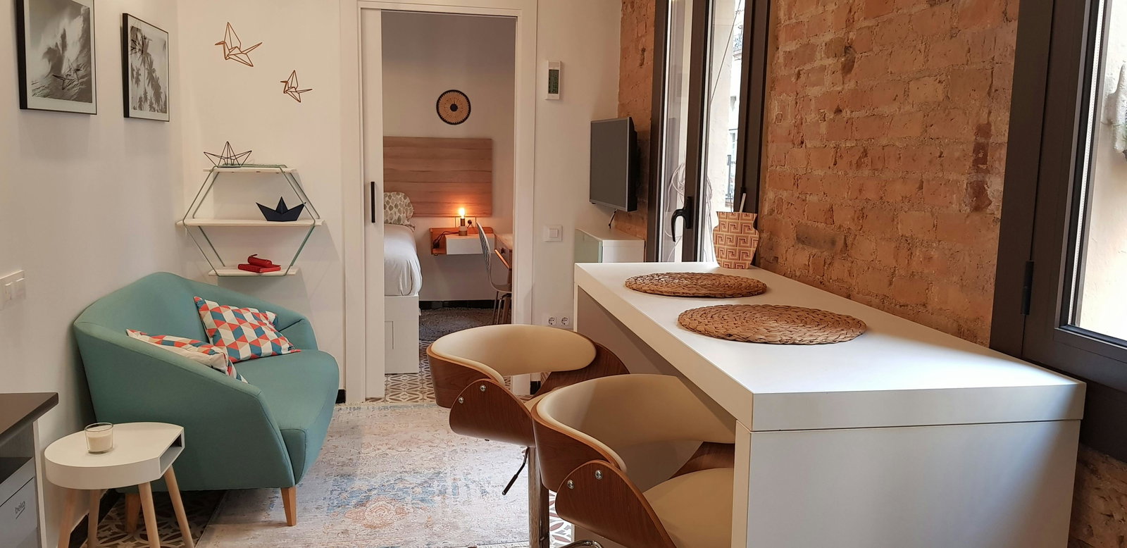 This is a view of the living room, featuring a light blue sofa with patterned cushions and a white side table with a candle. The room is tastefully decorated with minimalist furniture and an exposed brick wall.  A doorway leads into the bedroom, which features a queen-sized bed and a small desk.  The living room also includes a dining table with two chairs. There's also a window with a view of the city.  The overall ambiance of the room is bright and modern, with a touch of warmth.