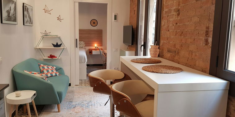 This is a view of the living room, featuring a light blue sofa with patterned cushions and a white side table with a candle. The room is tastefully decorated with minimalist furniture and an exposed brick wall. A doorway leads into the bedroom, which features a queen-sized bed and a small desk. The living room also includes a dining table with two chairs. There's also a window with a view of the city. The overall ambiance of the room is bright and modern, with a touch of warmth.