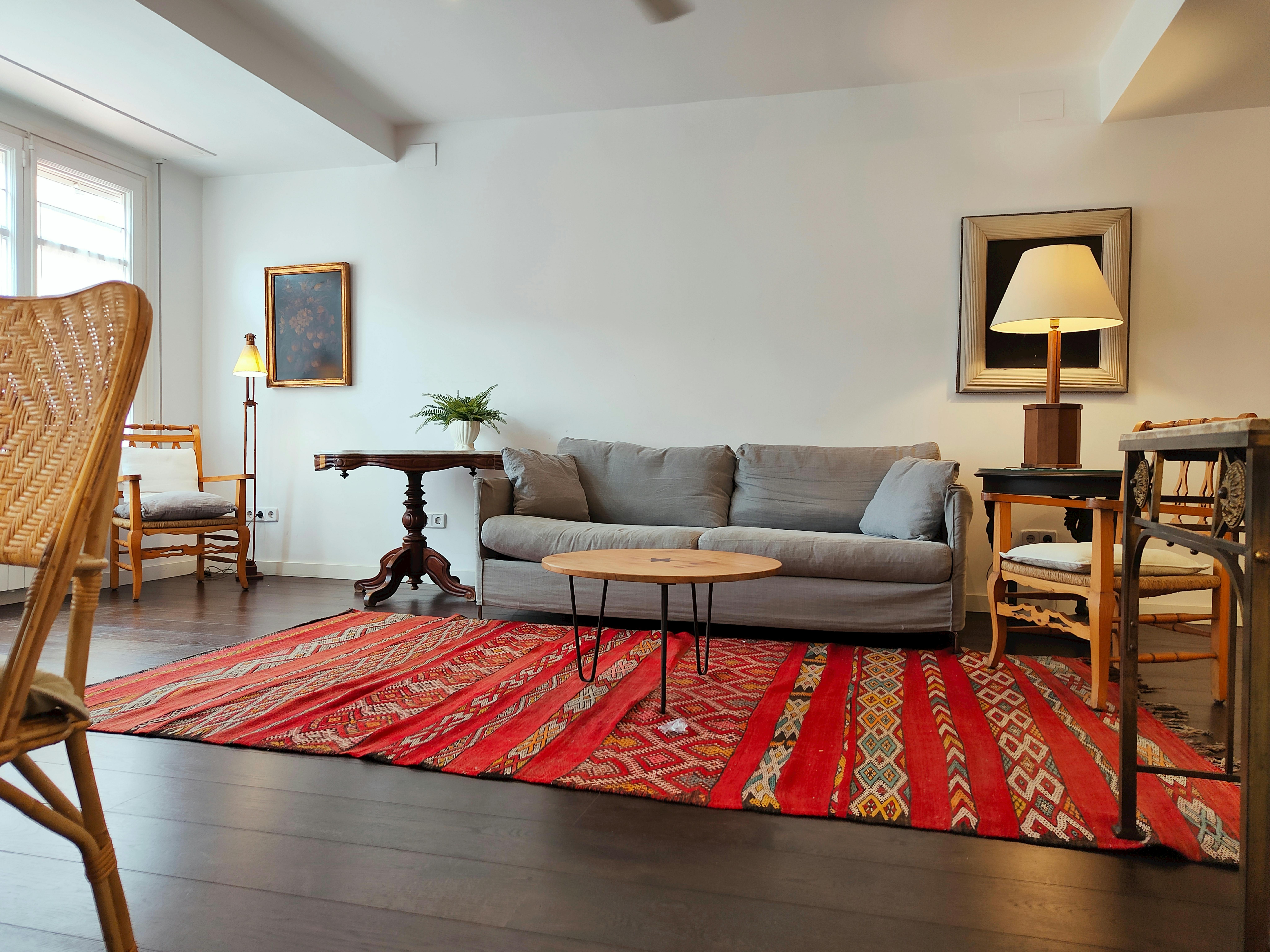 The living room features a comfortable grey sofa, a wooden armchair with cushions, a small side table, a floor lamp, and a large window with white curtains.  The sunlight streams through the window, illuminating the wooden floor and highlighting the intricate patterns of the colorful rug.  The space offers a welcoming and relaxed atmosphere.
