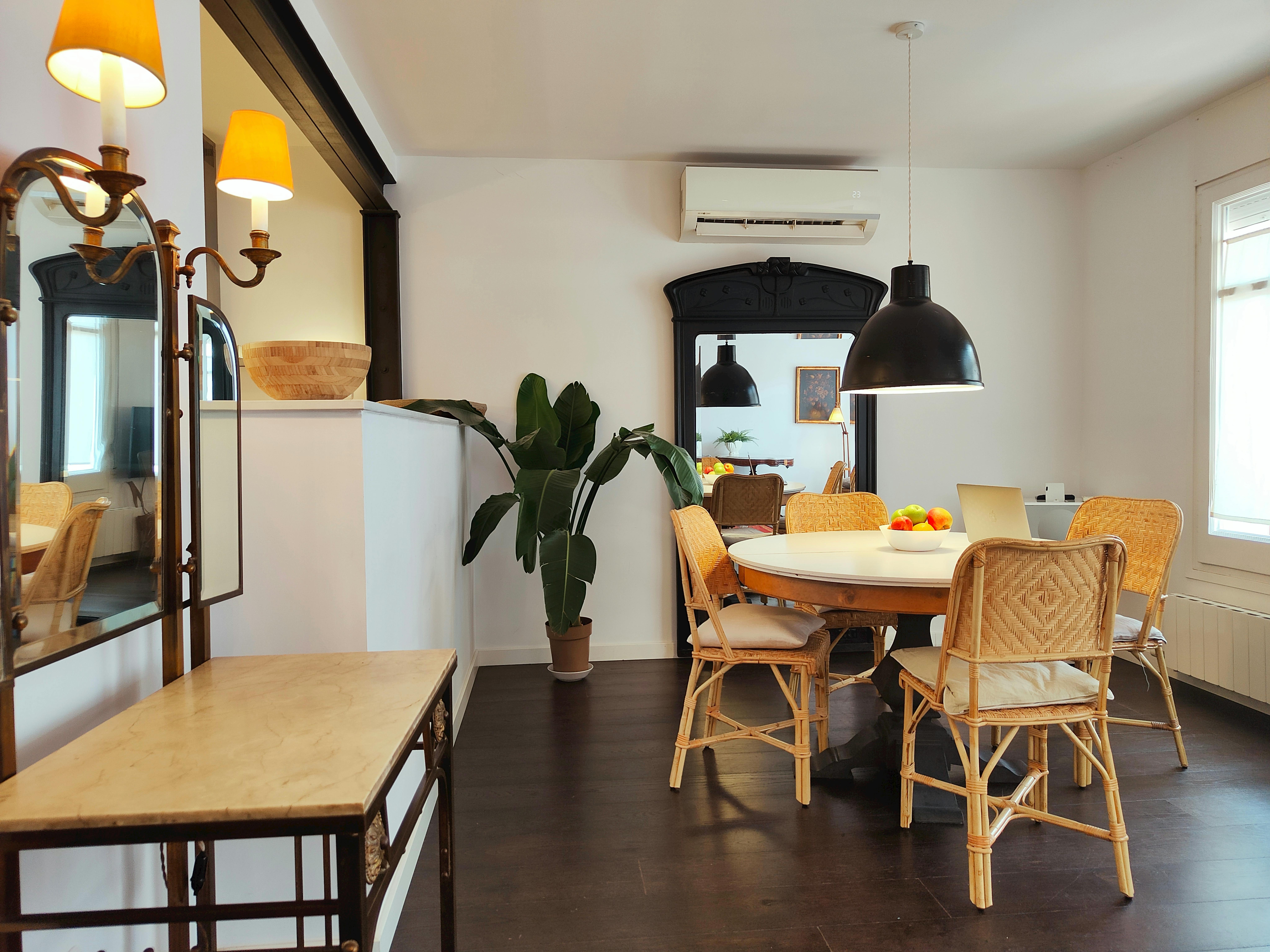 The dining area features a round wooden table surrounded by chairs with beige cushions. The room is bright and airy, with large windows allowing natural light to flood in.  A large mirror reflects the stylishly decorated dining area, providing a sense of depth and elegance. The dark wooden floors contribute to the overall warm and inviting ambiance.