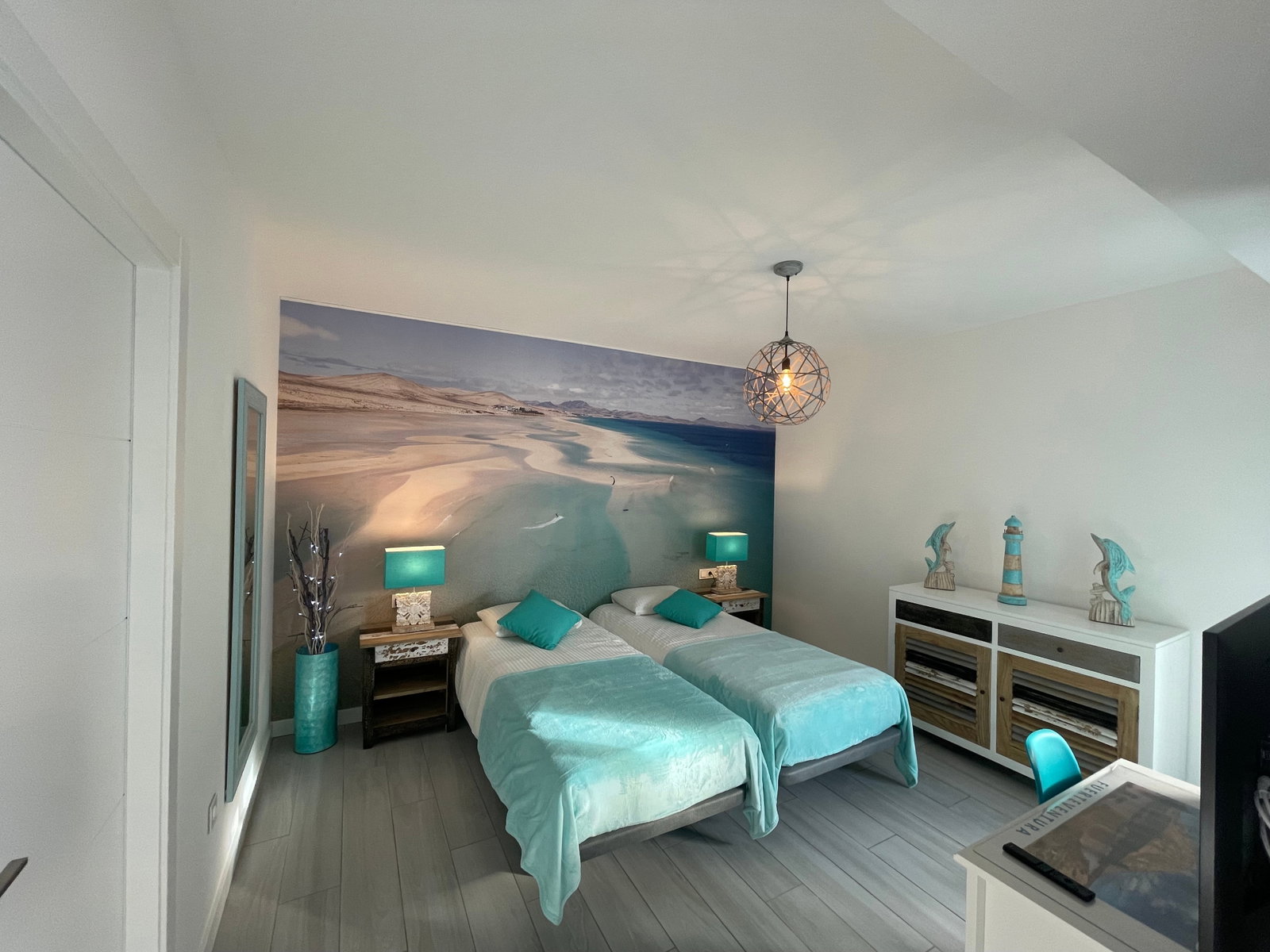 Cette chambre lumineuse vous plonge dans une ambiance balnéaire avec sa fresque murale évoquant les plages de Fuerteventura. Les touches de turquoise et le mobilier moderne créent un espace apaisant pour vous ressourcer.