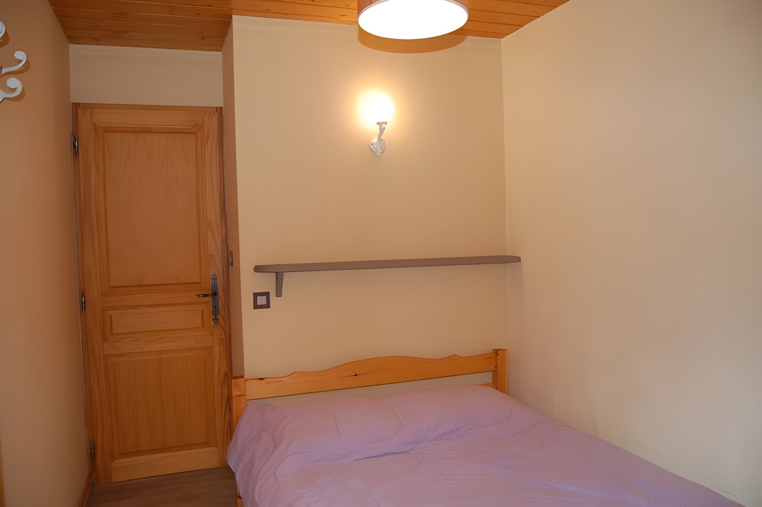 This is one of the three bedrooms in the apartment, featuring a double bed with a lavender-colored duvet. It is simply furnished with wooden floors, a small shelf, and a light fixture. The room is painted in a light beige color, creating a tranquil ambiance. A wooden door to the right leads to the hallway.
