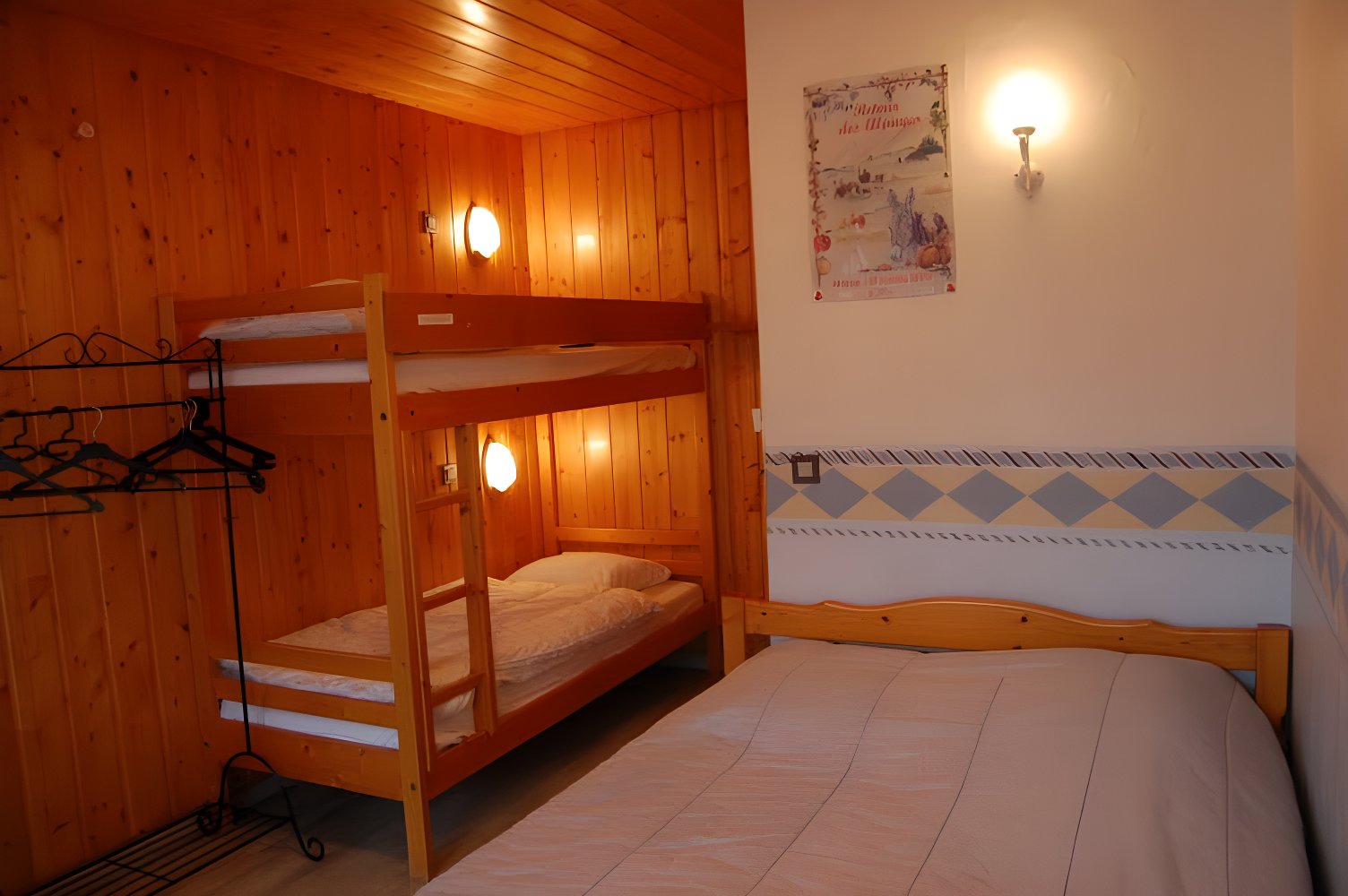 This cozy bedroom features a bunk bed with a single bed underneath, perfect for families or groups of friends. The room also includes a double bed with a white bedspread and a vintage poster on the wall, adding a touch of character to the space. The wooden walls and exposed beams create a warm and inviting atmosphere.
