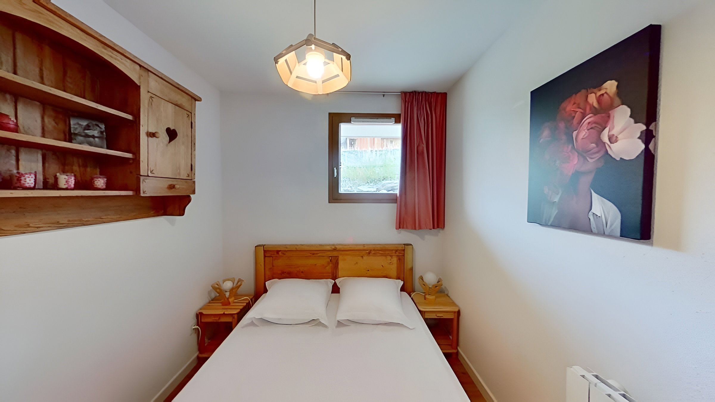 This inviting bedroom features a double bed with crisp white linens, wooden bedside tables with lamps, and a window with a view of the surrounding mountains. The room is decorated with a charming wooden headboard and a striking floral artwork above the heater, adding a touch of warmth and personality.  A rustic wooden shelf unit with a heart-shaped cut-out adds to the cozy ambiance of this space.