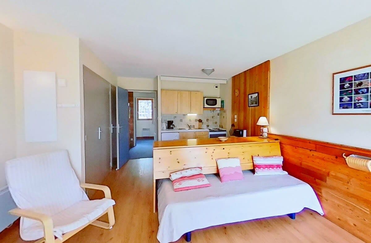 This inviting living area features a comfortable sofa bed, perfect for relaxing after a day of exploring the surrounding mountains. The sofa bed is situated near a large window, allowing for plenty of natural light and views of the picturesque alpine village. The space is further enhanced by a cozy armchair and a small table, ideal for enjoying a cup of coffee or reading a book. The open-plan kitchen, visible in the background, is fully equipped with modern appliances, ensuring that meal preparation is a breeze. The apartment's thoughtful design and warm, inviting atmosphere make it an ideal choice for couples and small families seeking a peaceful retreat in the heart