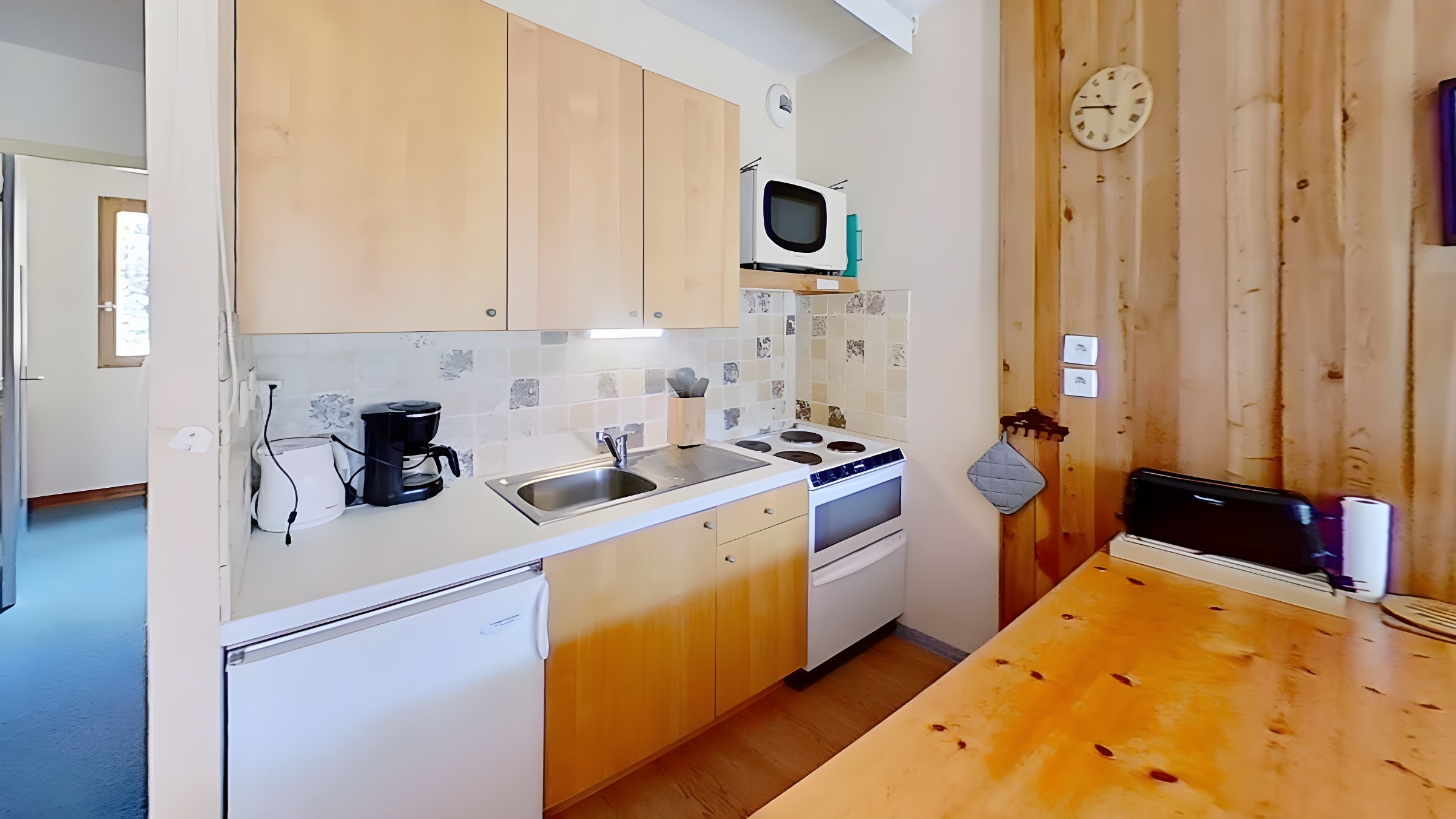 The well-equipped kitchen features a four-burner stovetop, a microwave, a refrigerator, and a dishwasher, perfect for preparing meals after a day of exploring the mountains. The charming tiled backsplash adds a touch of rustic elegance to the space. A coffee maker is provided for those who enjoy a morning brew. Natural light floods in through the window, illuminating the bright and airy space.  A wooden dining table is situated beside the kitchen, offering a comfortable spot to enjoy meals and unwind after a day of adventure.  A clock on the wall adds a touch of classic charm to the space, reminding guests of the tranquil pace