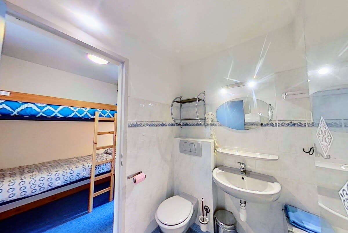 The bathroom features a white sink with a chrome faucet, a white toilet, and a large oval mirror above the sink. A small, open shelf is attached to the wall above the sink. To the left of the bathroom is a door that leads to a bunk bed alcove. The alcove features a wooden bunk bed with a blue and white patterned mattress and a wooden ladder.