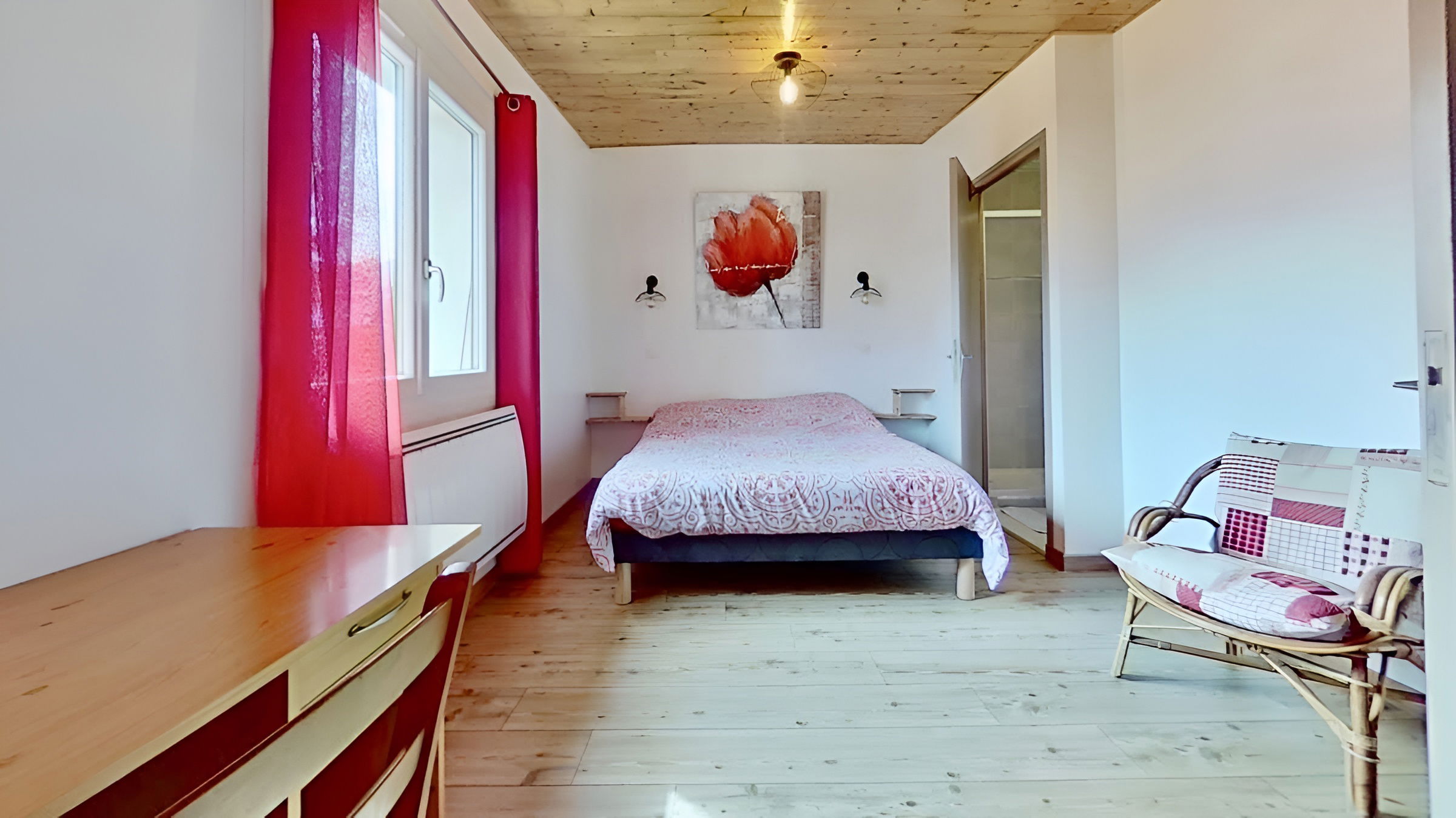 Inviting bedroom featuring a comfortable double bed with a patterned duvet. The light and airy space boasts a large window with bright red curtains, a charming rattan chair, and a simple desk. A cozy wood-paneled ceiling and a contemporary poppy painting add warmth and character to the room.