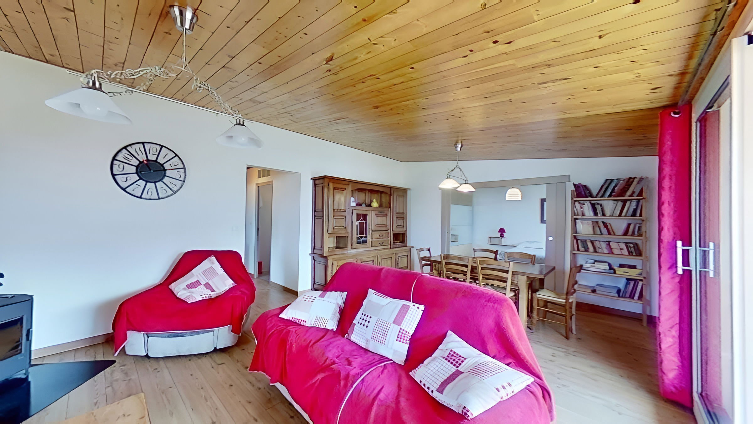 The spacious living room is furnished with a comfortable pink sofa, a matching armchair, and a wooden cabinet. The room features a wood-paneled ceiling and large windows providing plenty of natural light. An open doorway leads to the dining area, while a separate doorway provides access to the hallway. A decorative clock hangs on the wall and a wood-burning stove sits in the corner of the room.