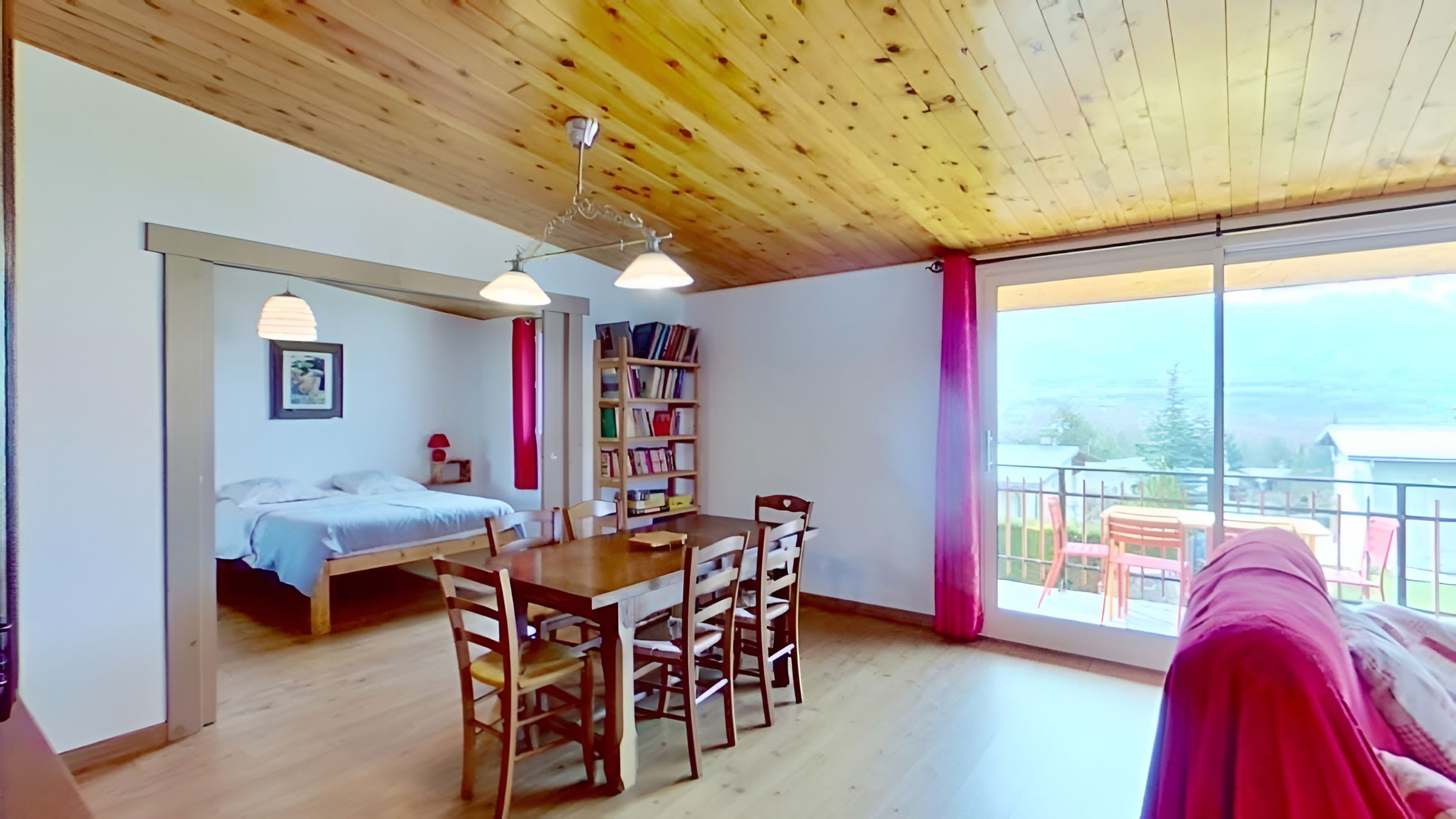 The spacious living room is furnished with a comfortable pink sofa, a matching armchair, and a wooden cabinet. The room features a wood-paneled ceiling and large windows providing plenty of natural light. An open doorway leads to the dining area, while a separate doorway provides access to the hallway. A decorative clock hangs on the wall and a wood-burning stove sits in the corner of the room.
