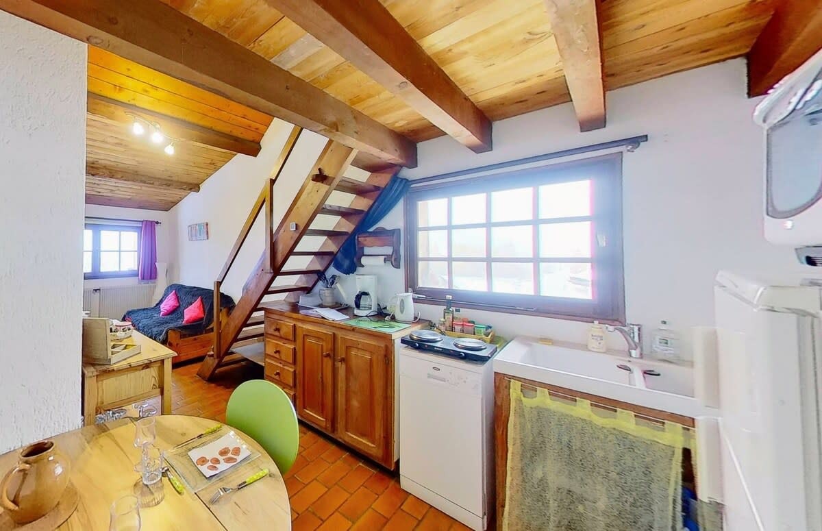 The studio's open-plan living space features a fully equipped kitchen with a dishwasher, a dining table for two, and a comfortable sofa bed. The wooden beams and exposed stonework give the space a charming, rustic feel. A staircase leads up to the mezzanine bedroom, providing a cozy and private sleeping area. The large window offers stunning views of the surrounding mountains.