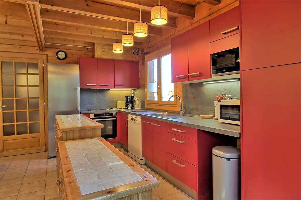 The chalet's fully equipped kitchen boasts modern appliances and a sleek design, featuring a breakfast bar with tiled countertop, a dishwasher, a microwave, and a bright red color scheme that adds a touch of vibrancy to the space. The kitchen offers a welcoming setting for preparing meals and enjoying a leisurely breakfast before heading out for a day of alpine adventures.