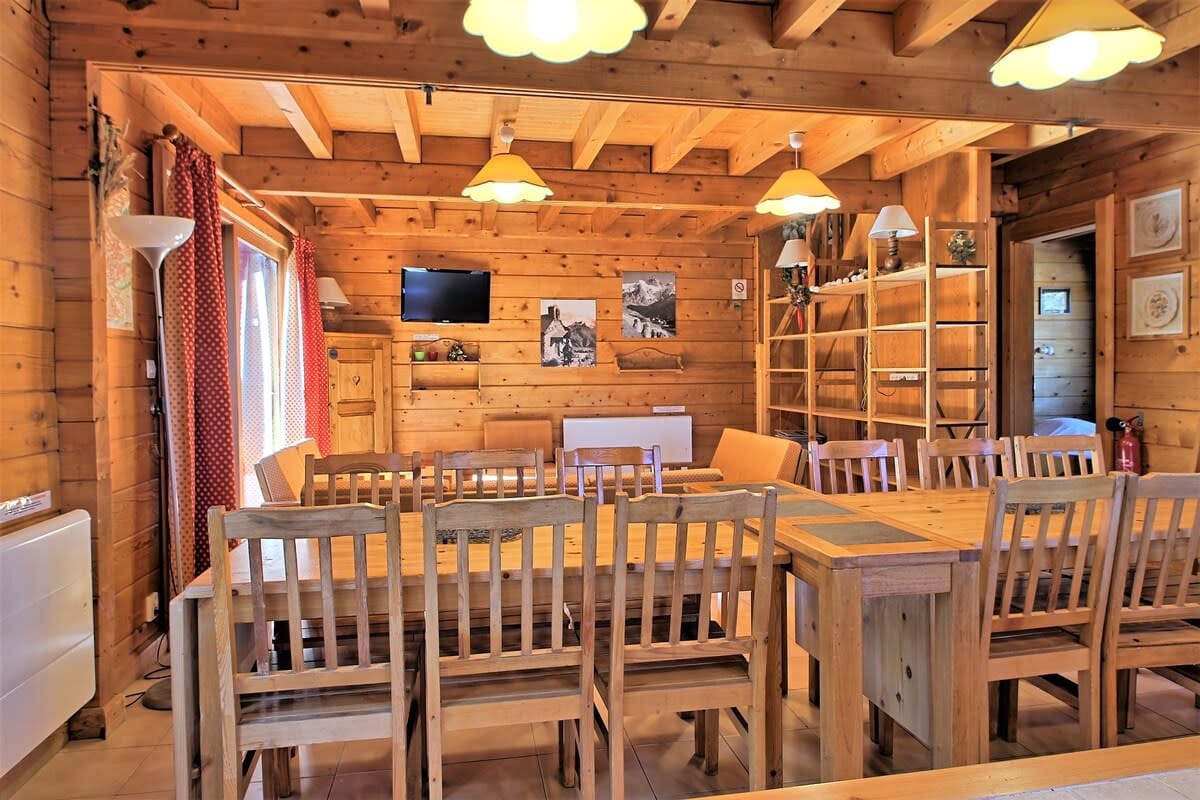 This spacious dining area features a large wooden table, perfect for gathering with loved ones.  The room is bathed in natural light from large windows and adorned with warm, wooden beams, creating a cozy and inviting atmosphere. The chalet's rustic charm is further enhanced by a wood-burning stove and a flat-screen television, providing both warmth and entertainment.  The well-equipped kitchen is visible in the background, offering a seamless transition between preparing meals and enjoying them with family and friends.  The open layout creates a sense of space and connection, making it ideal for both intimate dinners and lively gatherings.