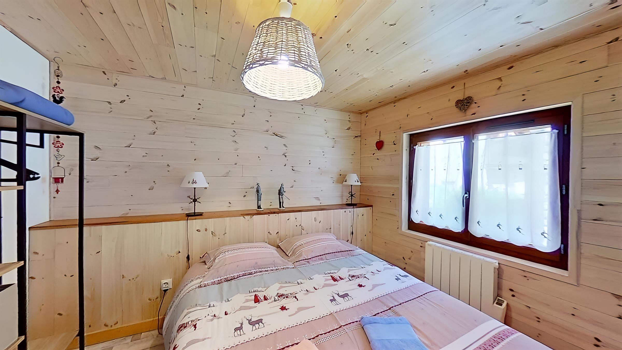 This cozy bedroom features a comfortable double bed with a charming winter-themed duvet cover and a warm wooden headboard. The room is bathed in natural light from a large window with sheer curtains, offering a glimpse of the surrounding mountain scenery.  The exposed wooden beams and walls add a rustic touch, creating a welcoming and inviting atmosphere.