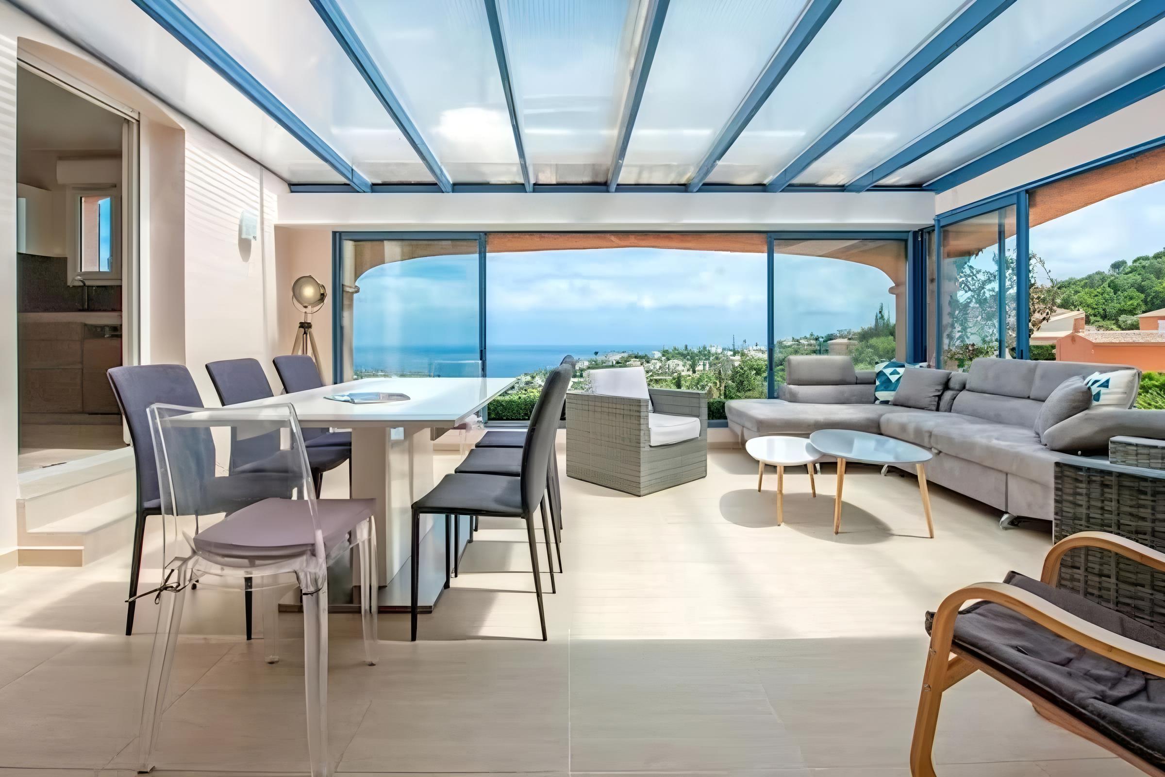 The spacious living area features a plush, grey sectional sofa, ideal for relaxing and enjoying the views.  Large windows offer an unobstructed view of the sparkling pool and surrounding greenery. The modern coffee tables provide ample space for drinks and snacks while taking in the serene atmosphere.  The villa's unique charm is further enhanced by the elegant wicker armchair in the corner, adding a touch of sophistication to the space.