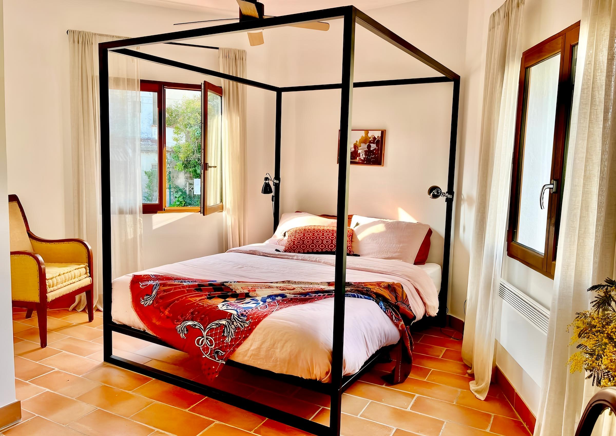 This bedroom showcases a comfortable double bed with a black metal canopy frame, bathed in natural light from large windows with flowing white curtains. A vibrant throw adds color to the minimalist decor, while a cozy armchair near the window offers tranquil views of the surroundings.