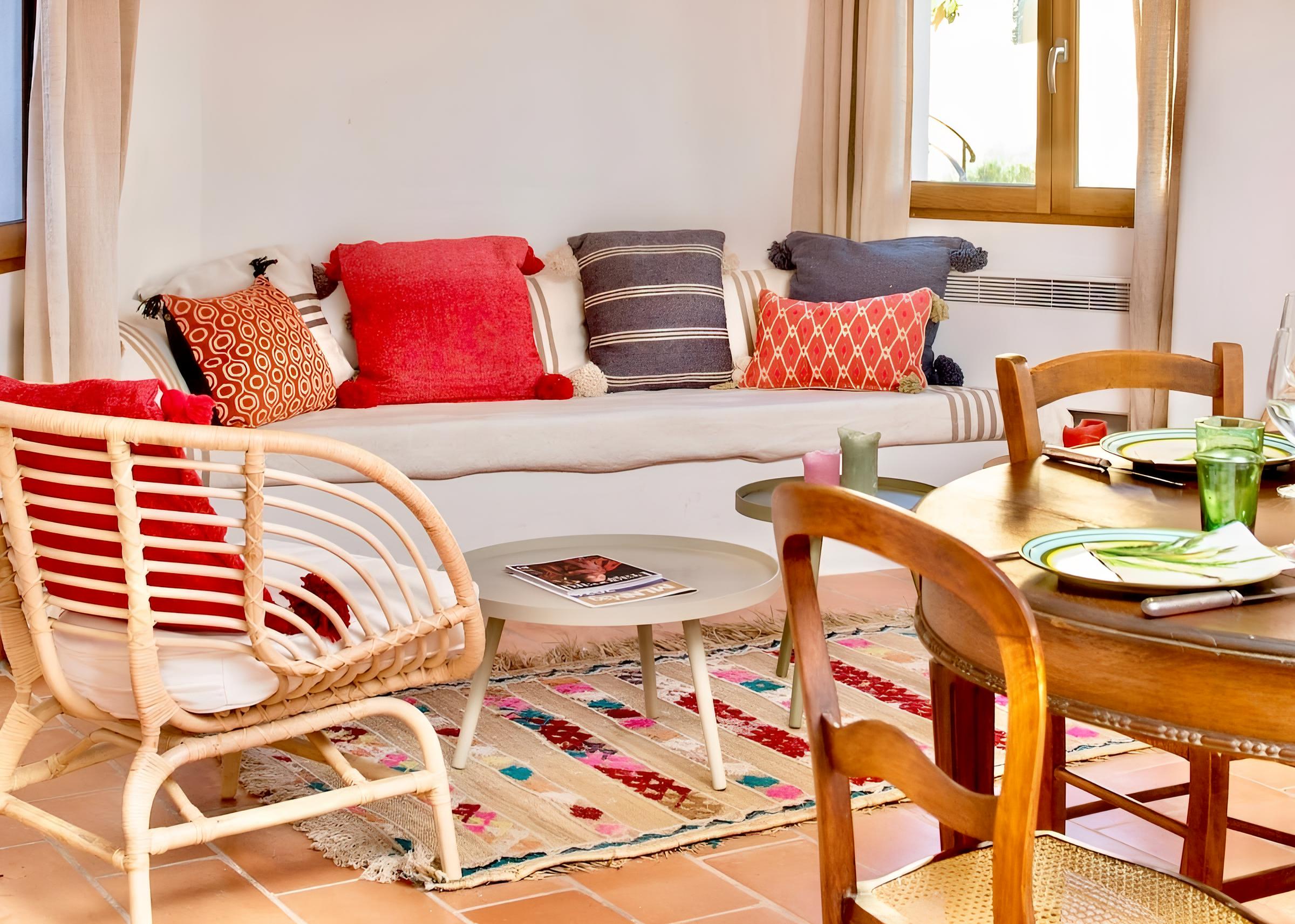 Spacious living area with a colorful bench, wicker armchair, Moroccan rug, and round coffee table. A wooden dining table and large windows provide a warm, welcoming atmosphere.
