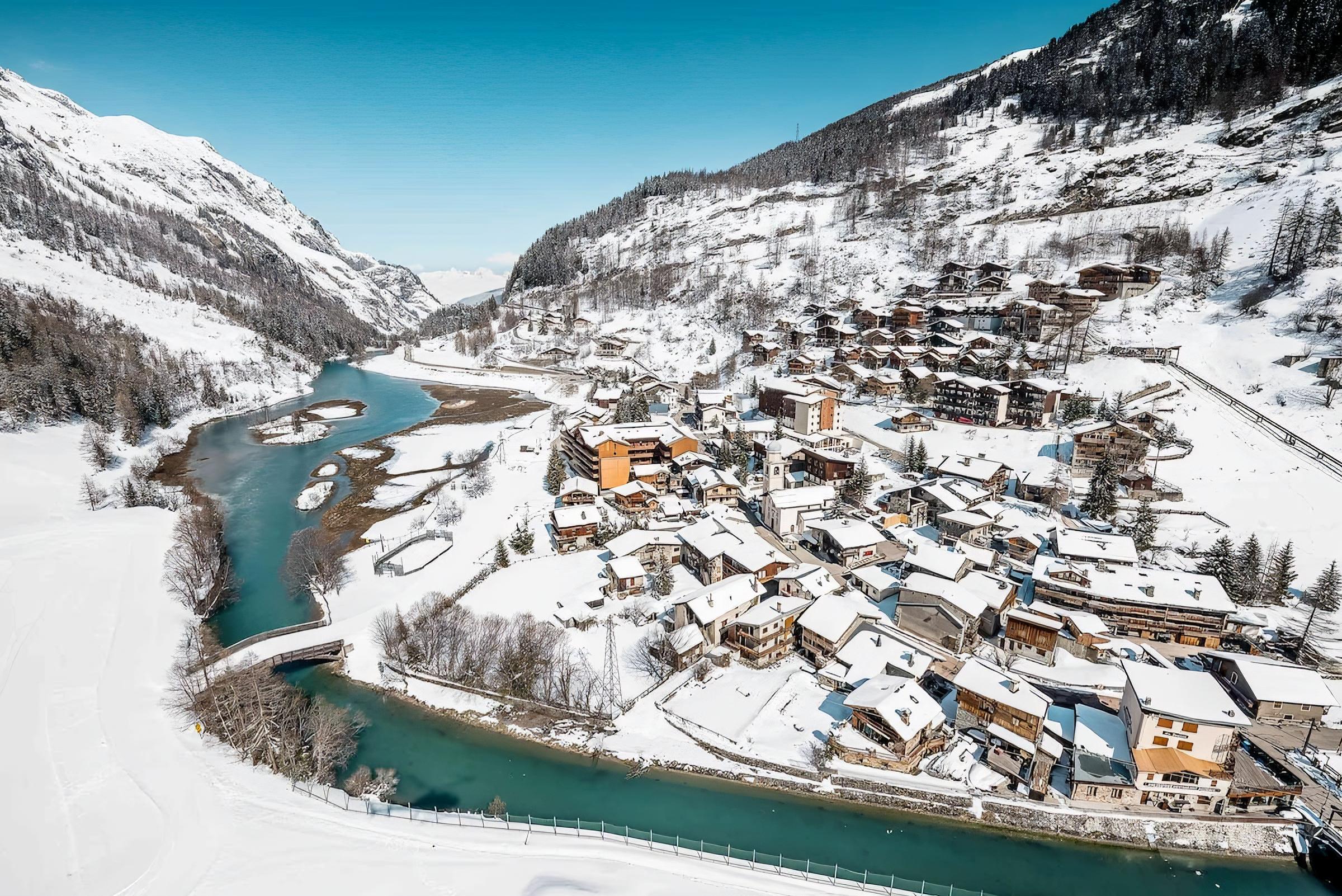 Enjoy breathtaking views of the charming village of Tignes-les-Brevières, nestled in the French Alps. The apartment is conveniently located just steps away from the village center, providing easy access to shops, restaurants, and après-ski activities. This panoramic view showcases the picturesque setting of the apartment, with snow-capped mountains, a winding river, and traditional alpine architecture.