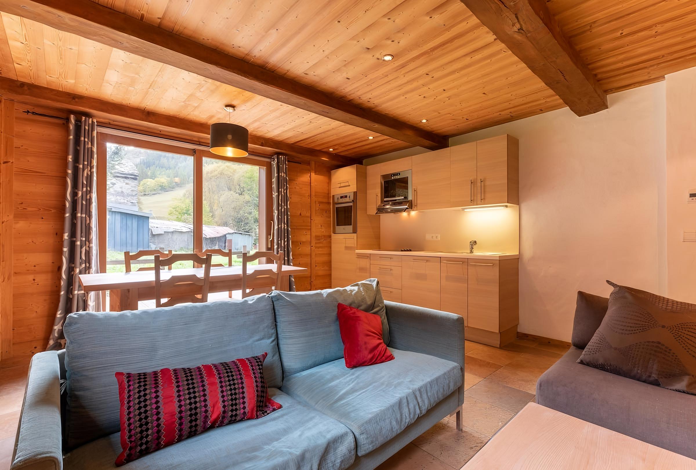 The open-plan living space features a comfortable blue sofa with colorful throw pillows, offering a cozy spot to relax after a day on the slopes. The wood-paneled walls and ceiling add a touch of alpine charm, while the large window offers views of the surrounding mountains and greenery. The fully equipped kitchen is visible in the background, providing everything needed to prepare meals during your stay.