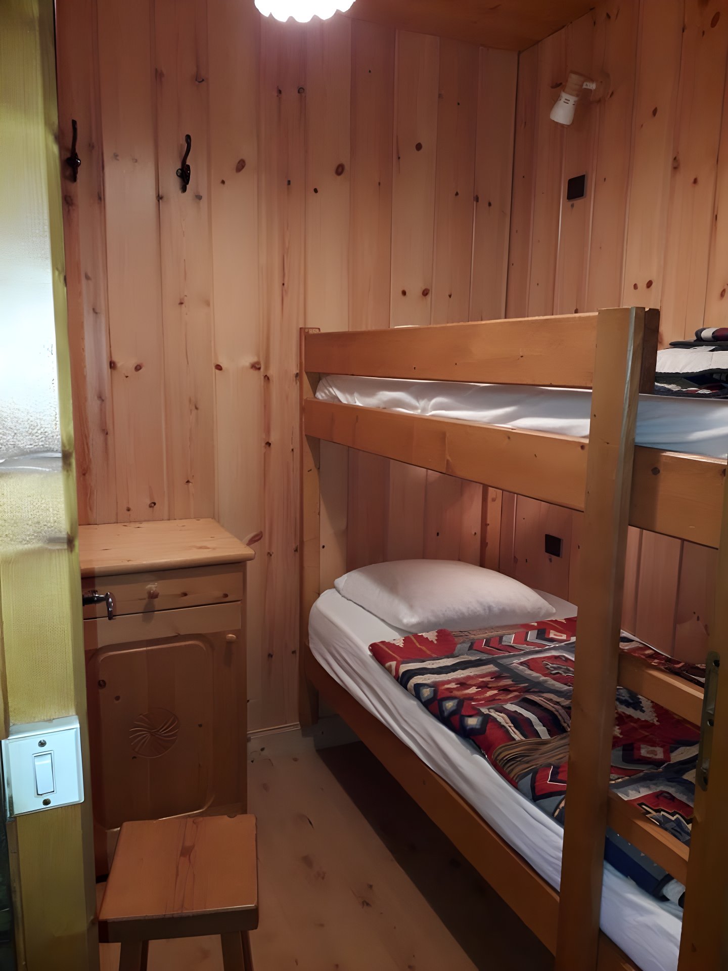 The cabin bedroom features two 90cm bunk beds with a window, creating a cozy and inviting space. The wooden bunk beds,  adorned with a colorful patterned blanket,  are sure to provide a restful sleep after a day exploring the mountains. The room's rustic charm is enhanced by the wooden walls and the small wooden stool next to the compact wooden cabinet.