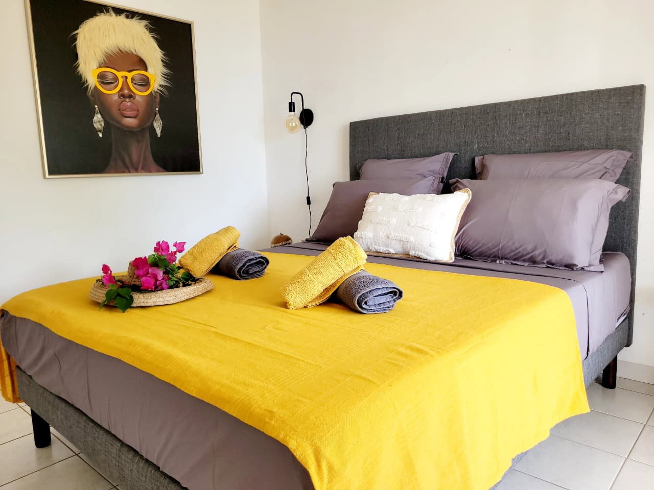 Chambre lumineuse & climatisée avec lit Queen Size confortable. Elle offre un mix d'oreillers carrés et rectangulaires pour un soutien optimal. Profitez de l'accès direct à la terrasse pour des réveils en douceur face au jardin tropical.