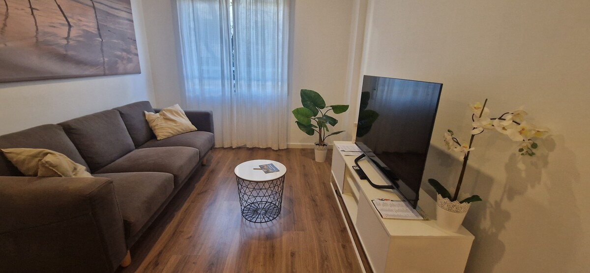 Acogedor salón-comedor con amplias ventanas y vistas a la calle, aire acondicionado, mesa para cuatro y un sofá ideal para descansar o ver la televisión.