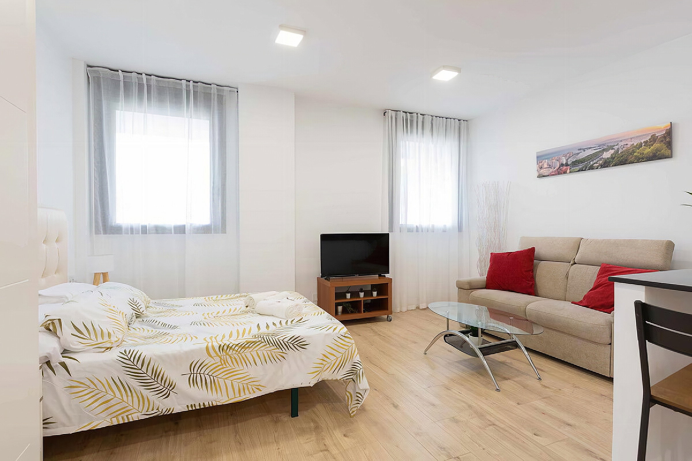 A connecting room with a bedroom. A double bed with a sofa. A coffee table, a dresser, and a TV, as well as two windows with long curtains.