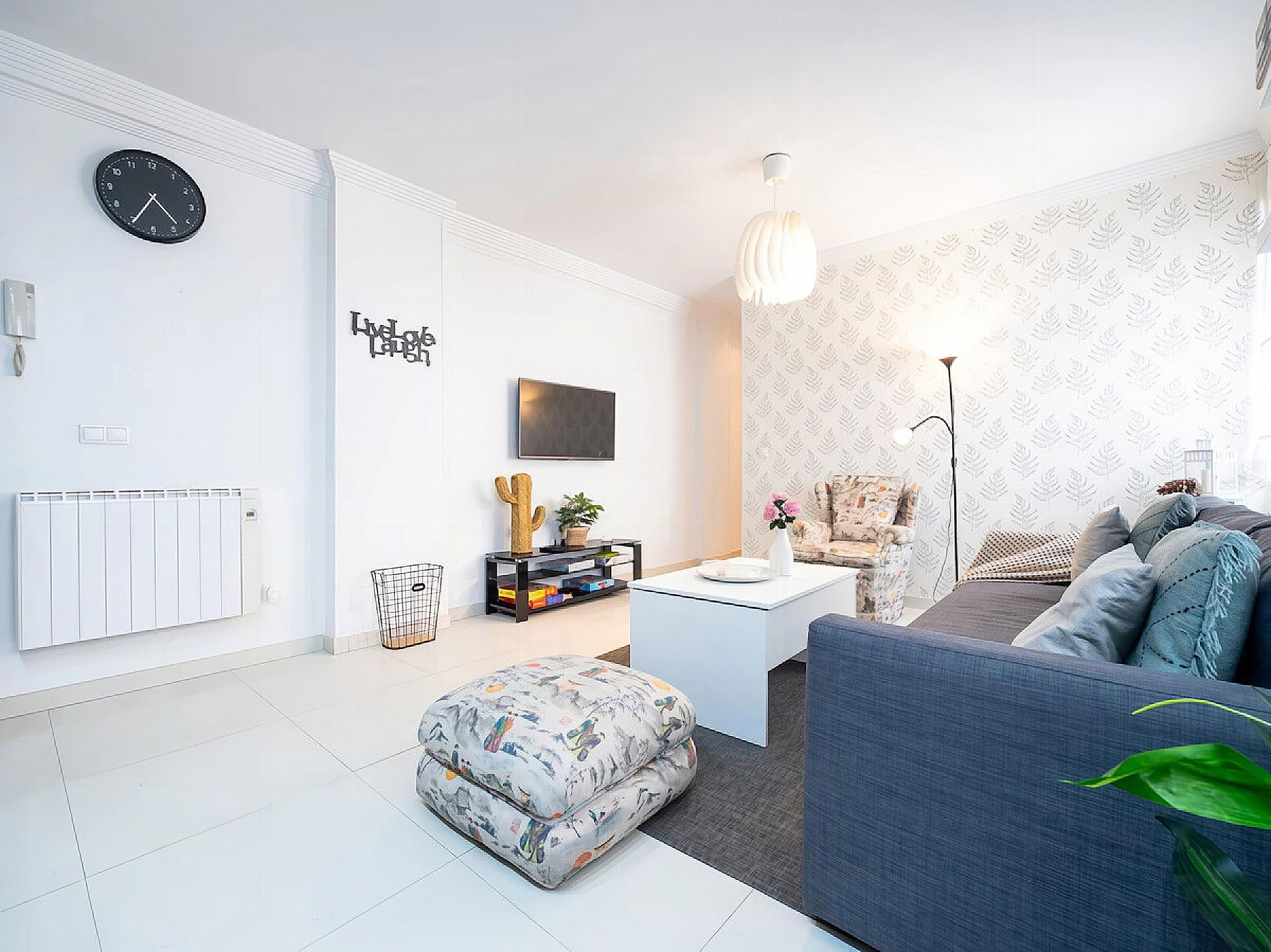 Communal lounge with TV, sofa, coffee table, armchair, ottoman, small chest of drawers with plants on top, wallpaper on the wall.